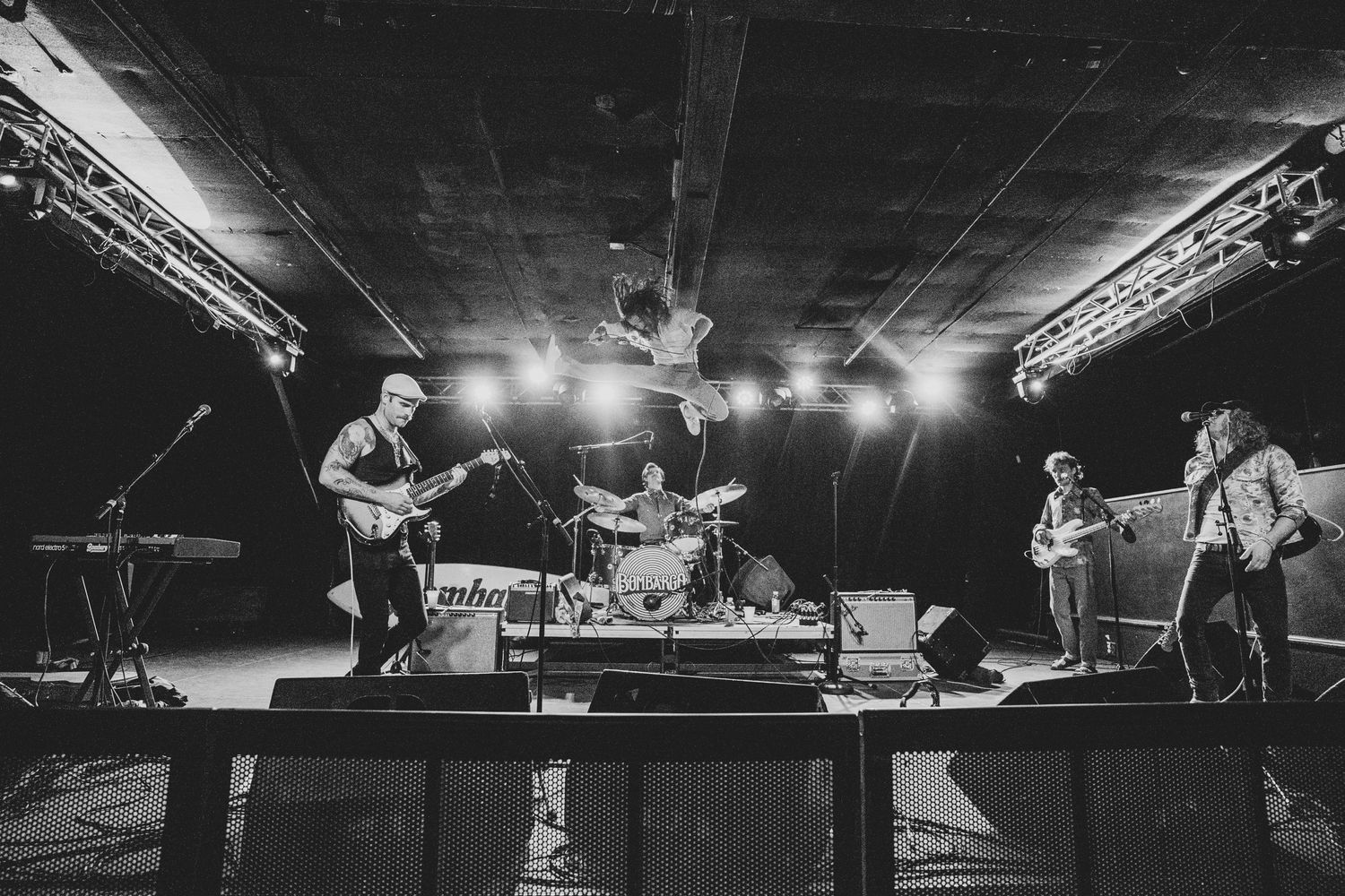 Musicians play instruments on a dimly lit stage with overhead lighting and sound equipment.