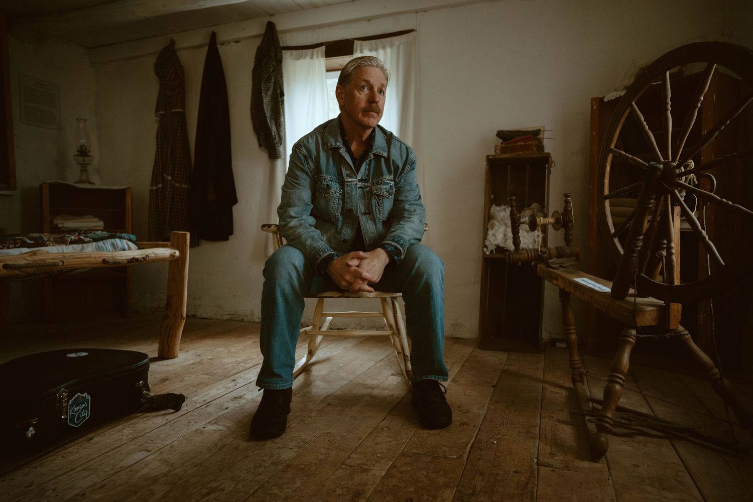 A person sits in contemplative pose on a wooden chair in a rustic room with spinning wheel and natural lighting.