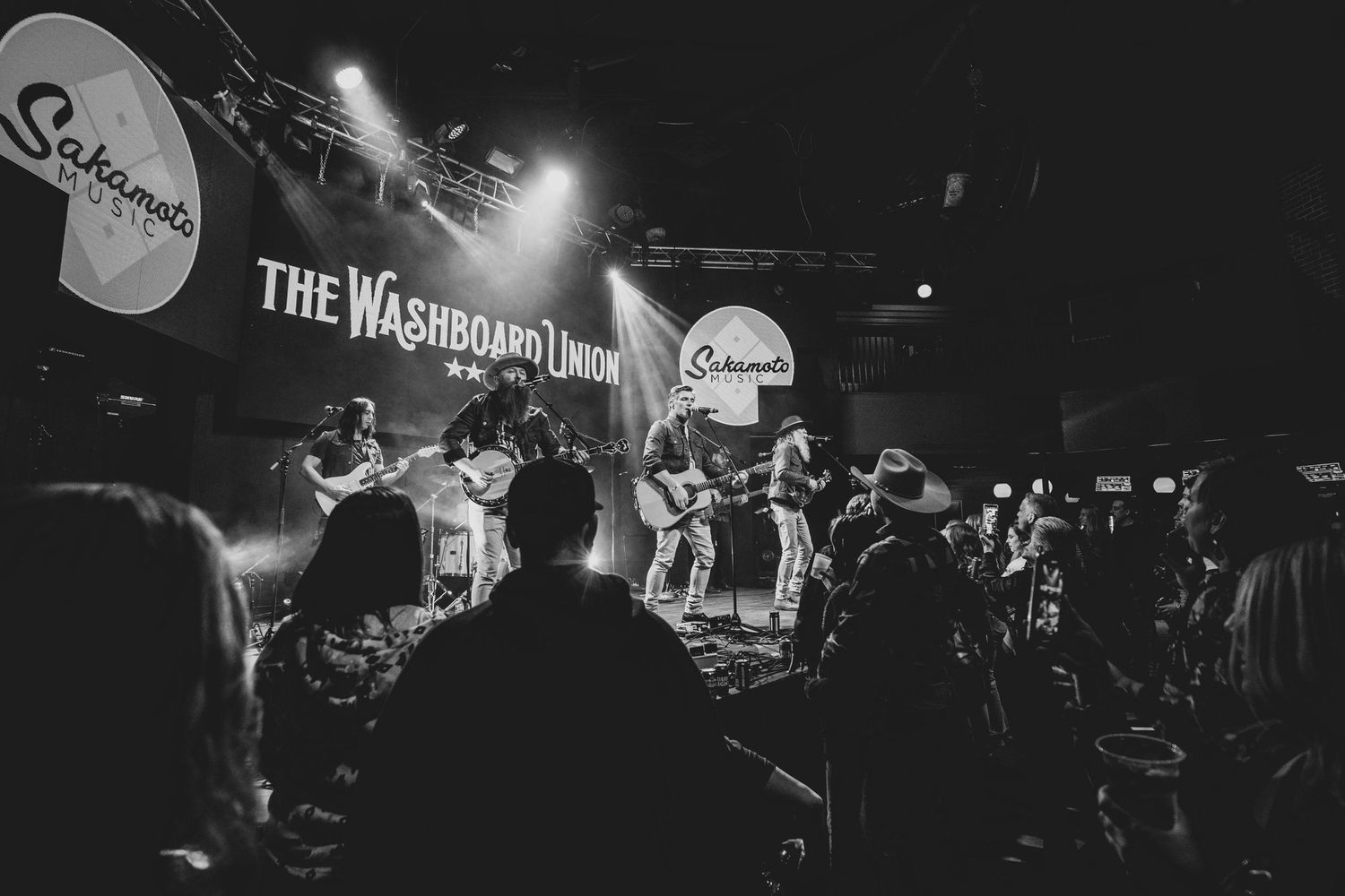 Wide shot of band performing on stage with crowd silhouettes in foreground.
