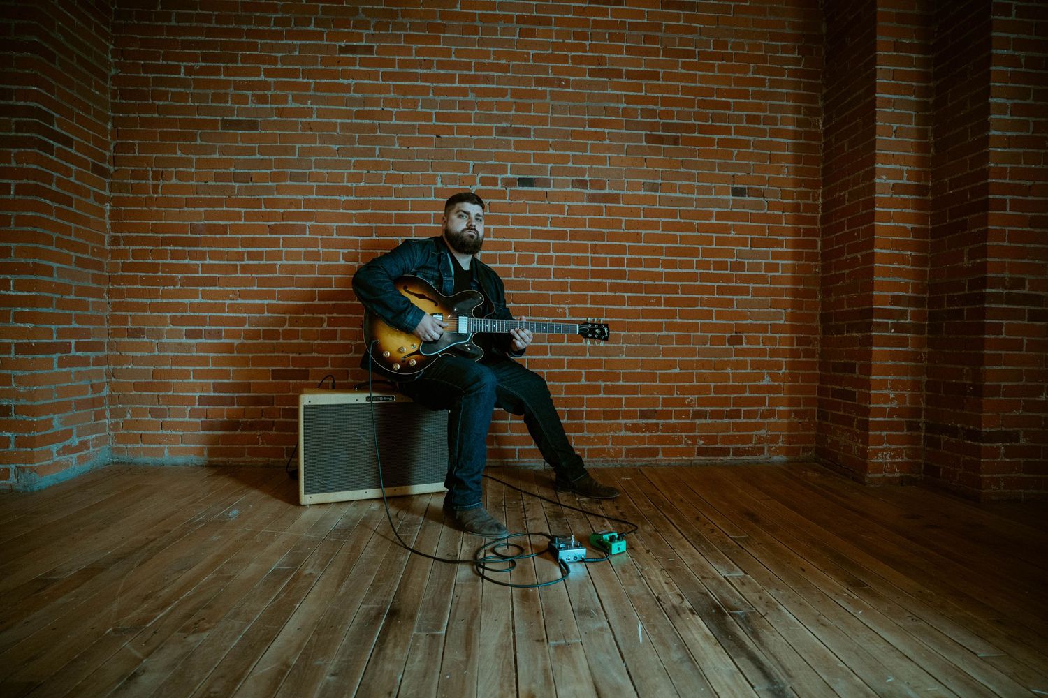 Dylan Farrell performs with electric guitar in an empty brick-walled room with pedals on floor.