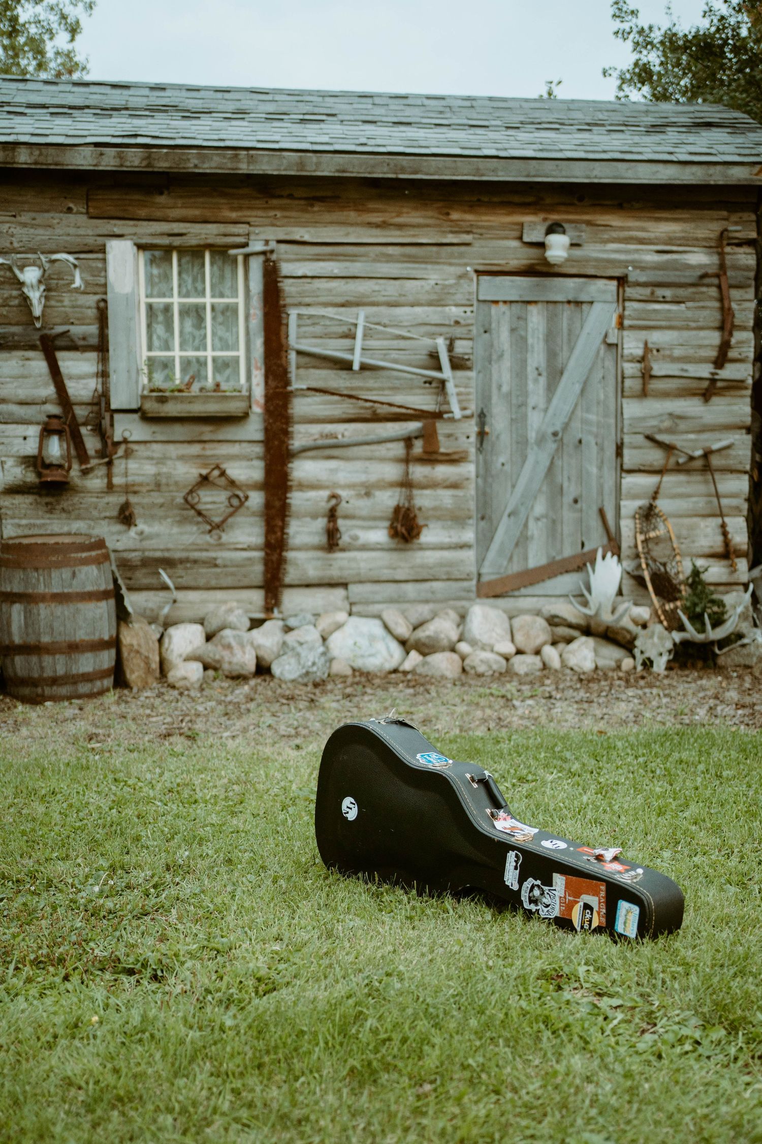 Guitar case lying on grass in front of weathered wooden cabin with tools hanging on exterior wall for Tim Isberg's album artwork.