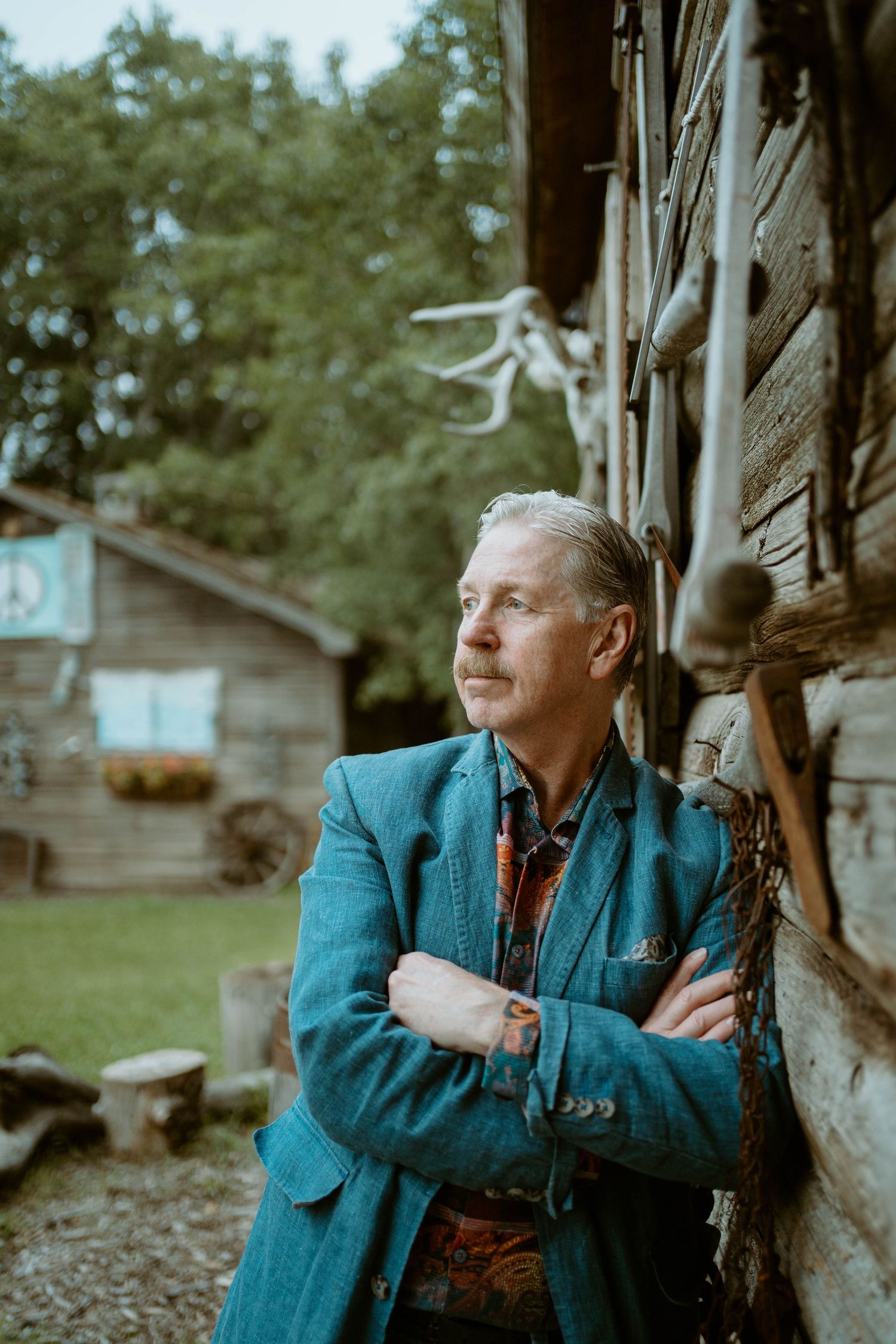 Tim Isberg in turquoise blazer gazes thoughtfully while leaning against rustic log cabin wall.