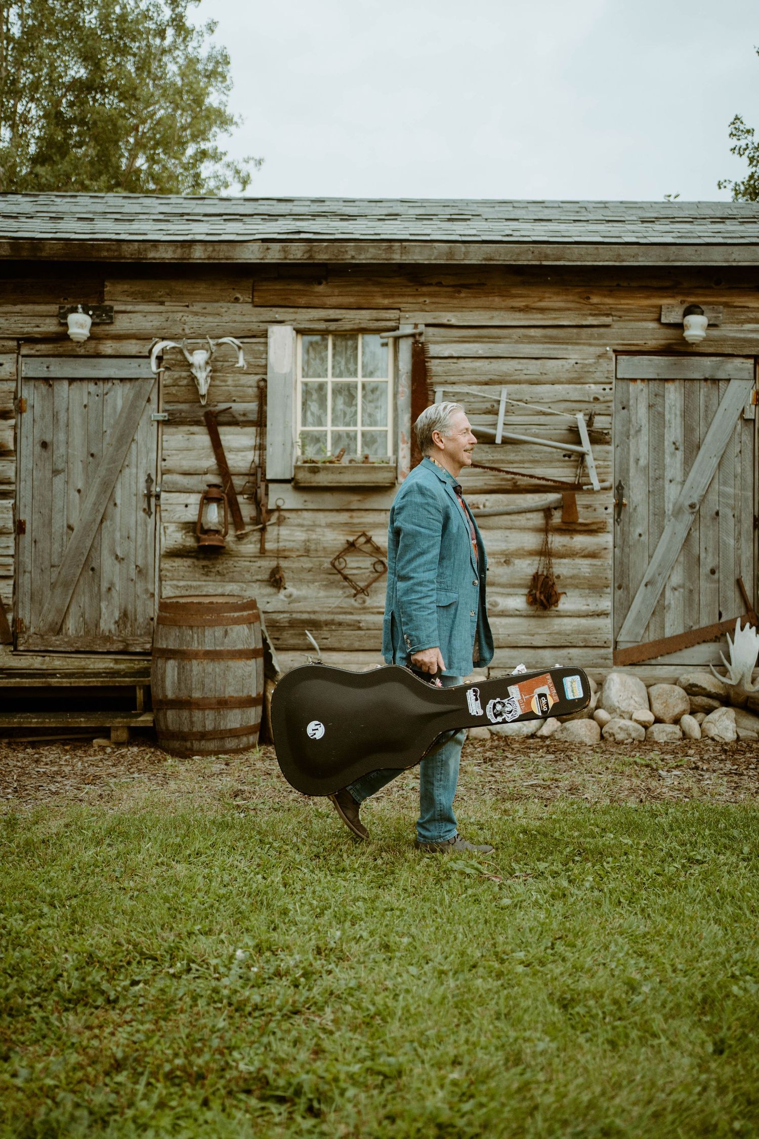 Tim Isberg in blue jacket carrying guitar case outside rustic log cabin with vintage tools on wall.