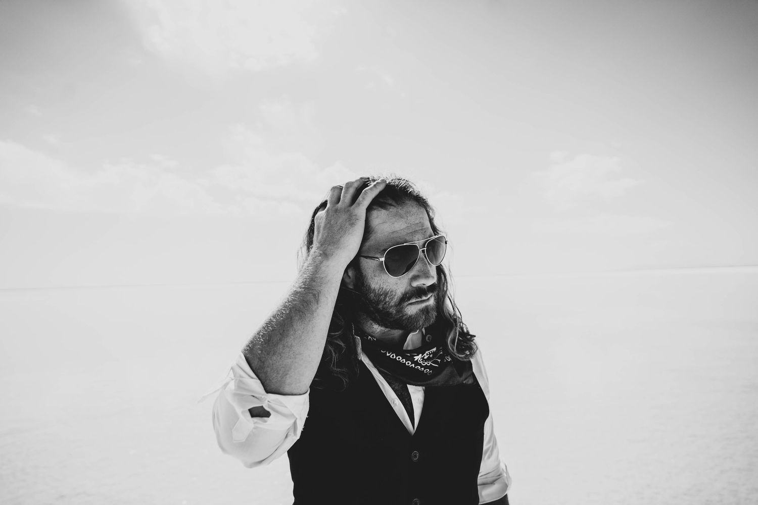 Black and white candid shot of person in vest and sunglasses with hand on head.