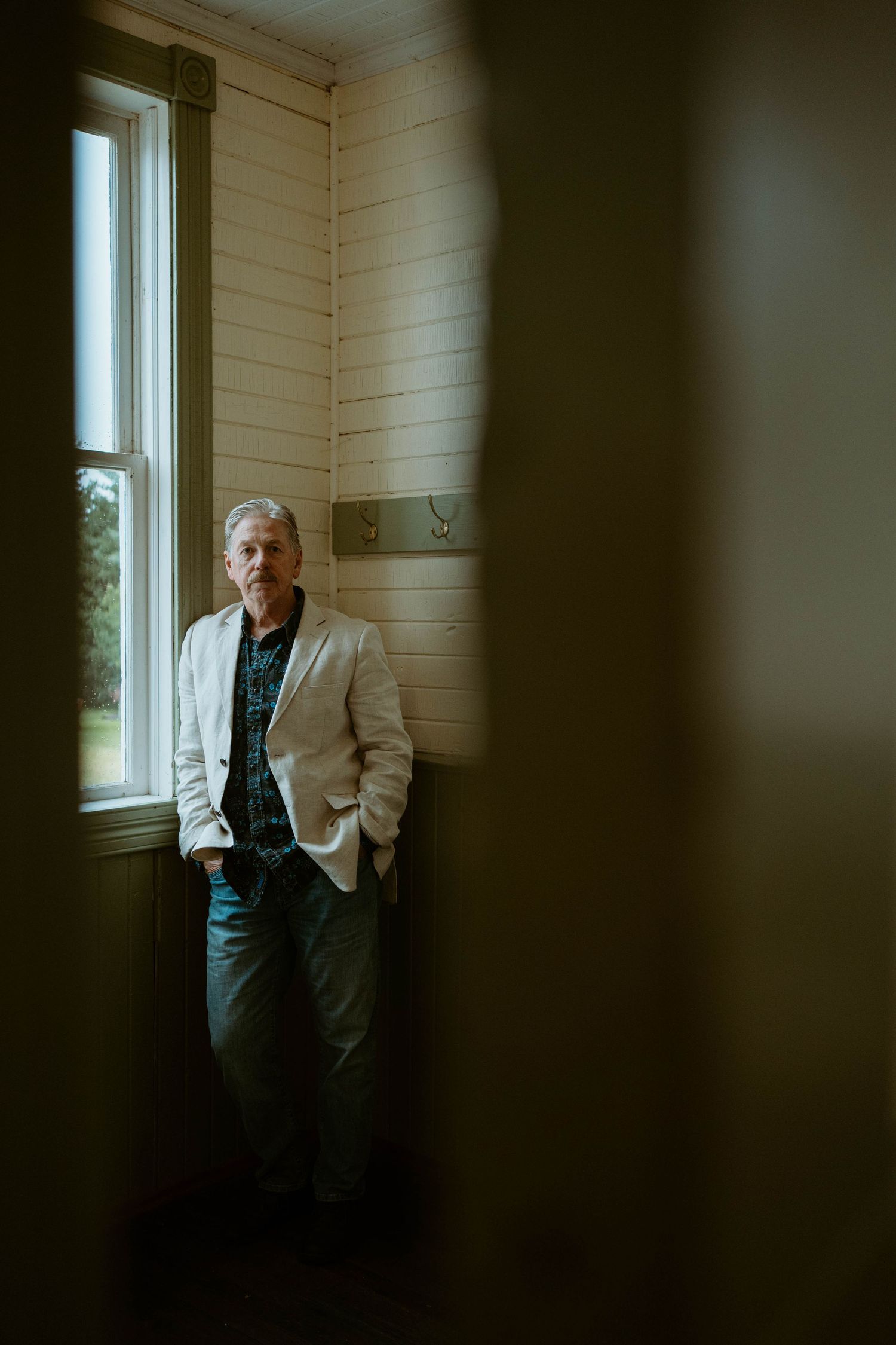 A figure stands in contemplative pose by a bright window in a minimalist white-walled room.