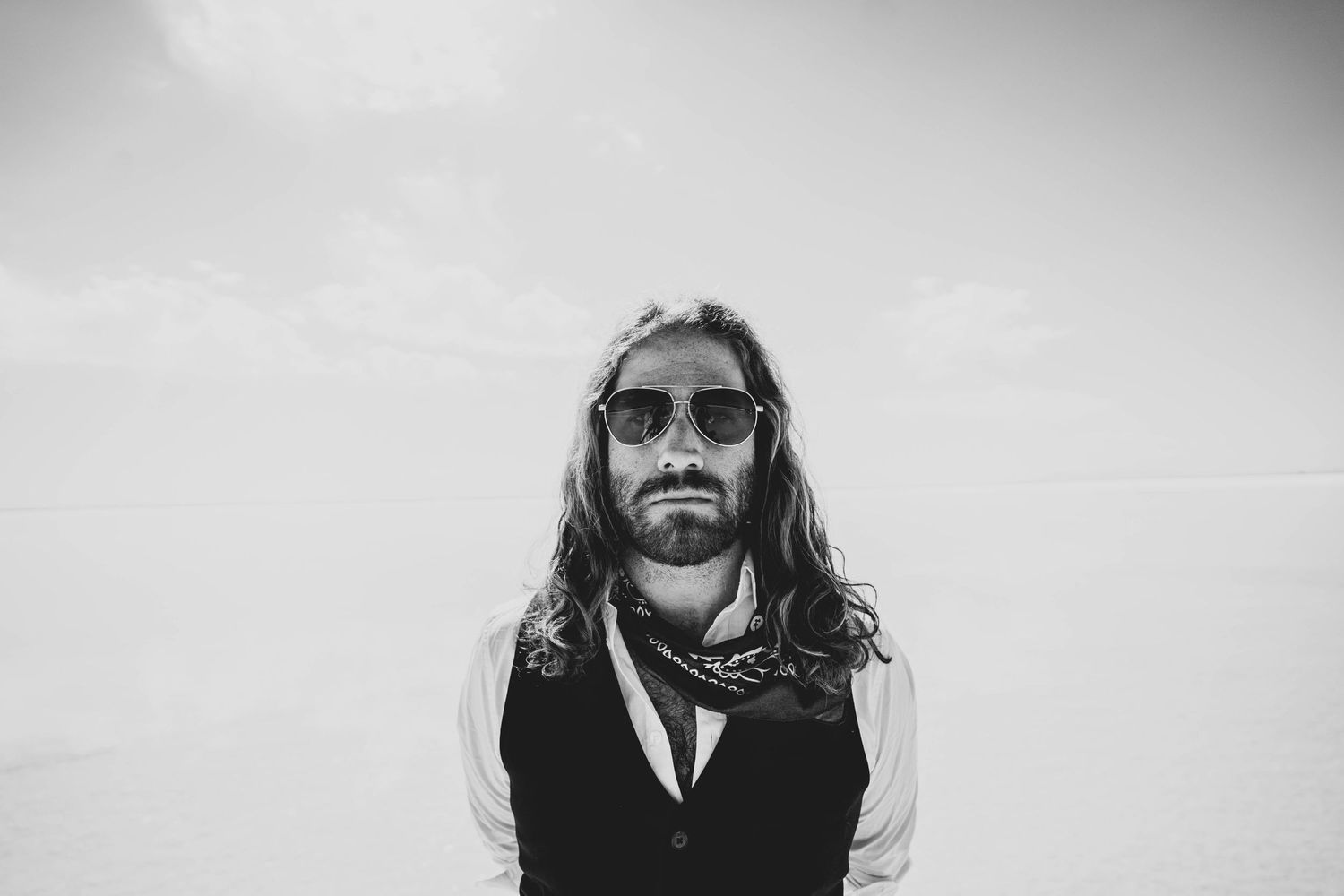 Black and white portrait of person wearing sunglasses and a vest with long hair smiling against plain background.