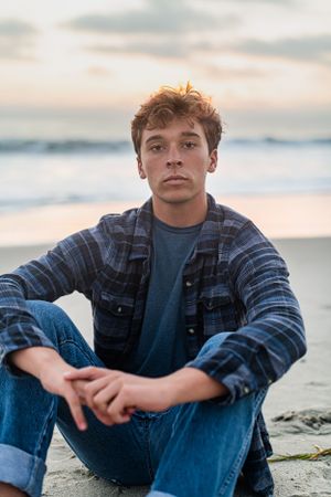 Teen boy sitting on beach at dusk in plaid shirt and jeans.