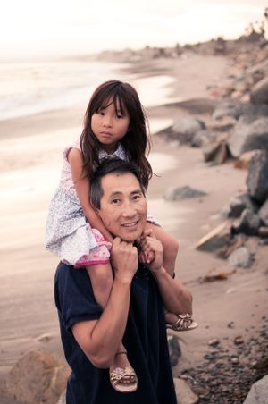 Smiling father carrying young daughter on his shoulders on a rocky beach at sunset.