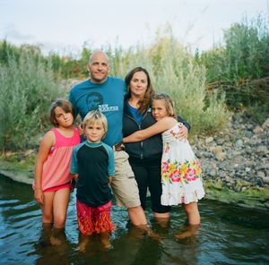 Family of five standing together in a shallow river surrounded by lush green vegetation outdoors.