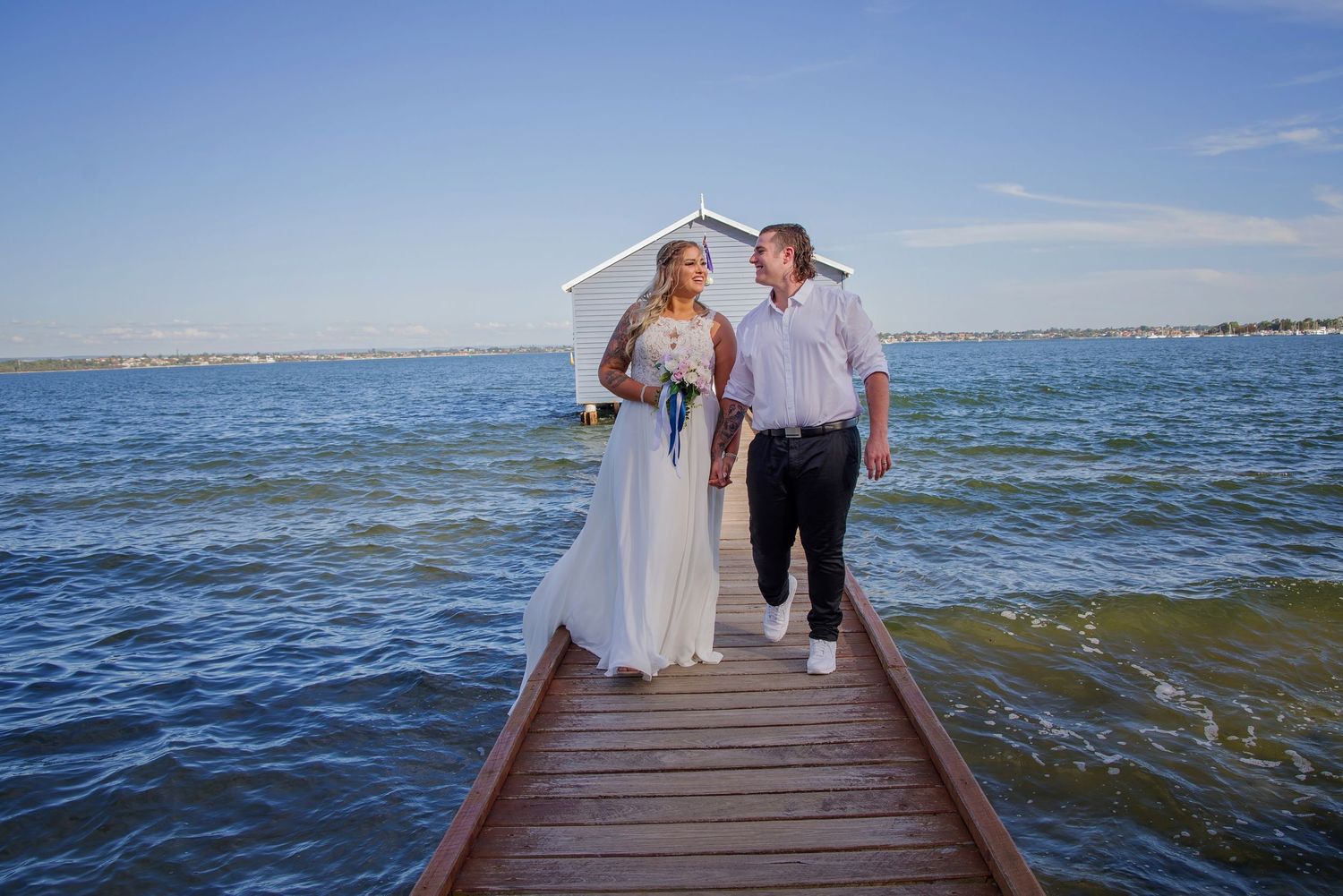 bride and groom walking on bridge and smiling at each other blue boat house behind them