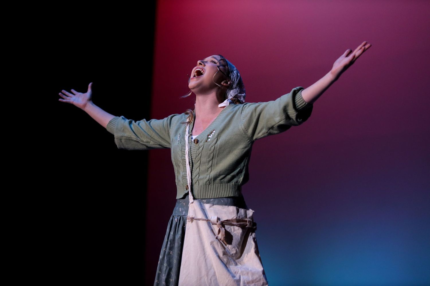 Dramatic stage performance with performer in vintage dress with arms spread under colored lighting.
