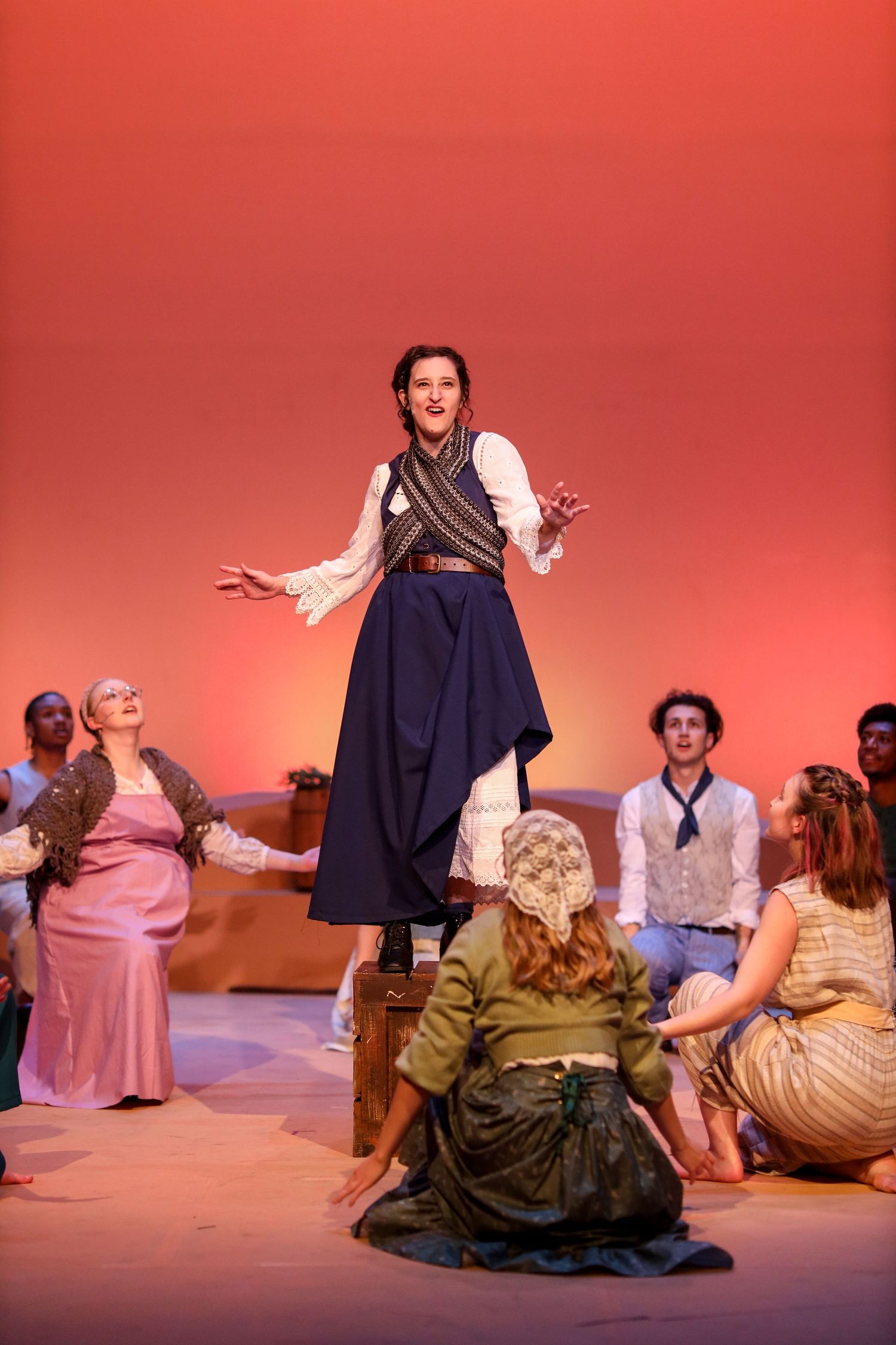 Theater performer in period costume addressing the audience while others sit around the stage.