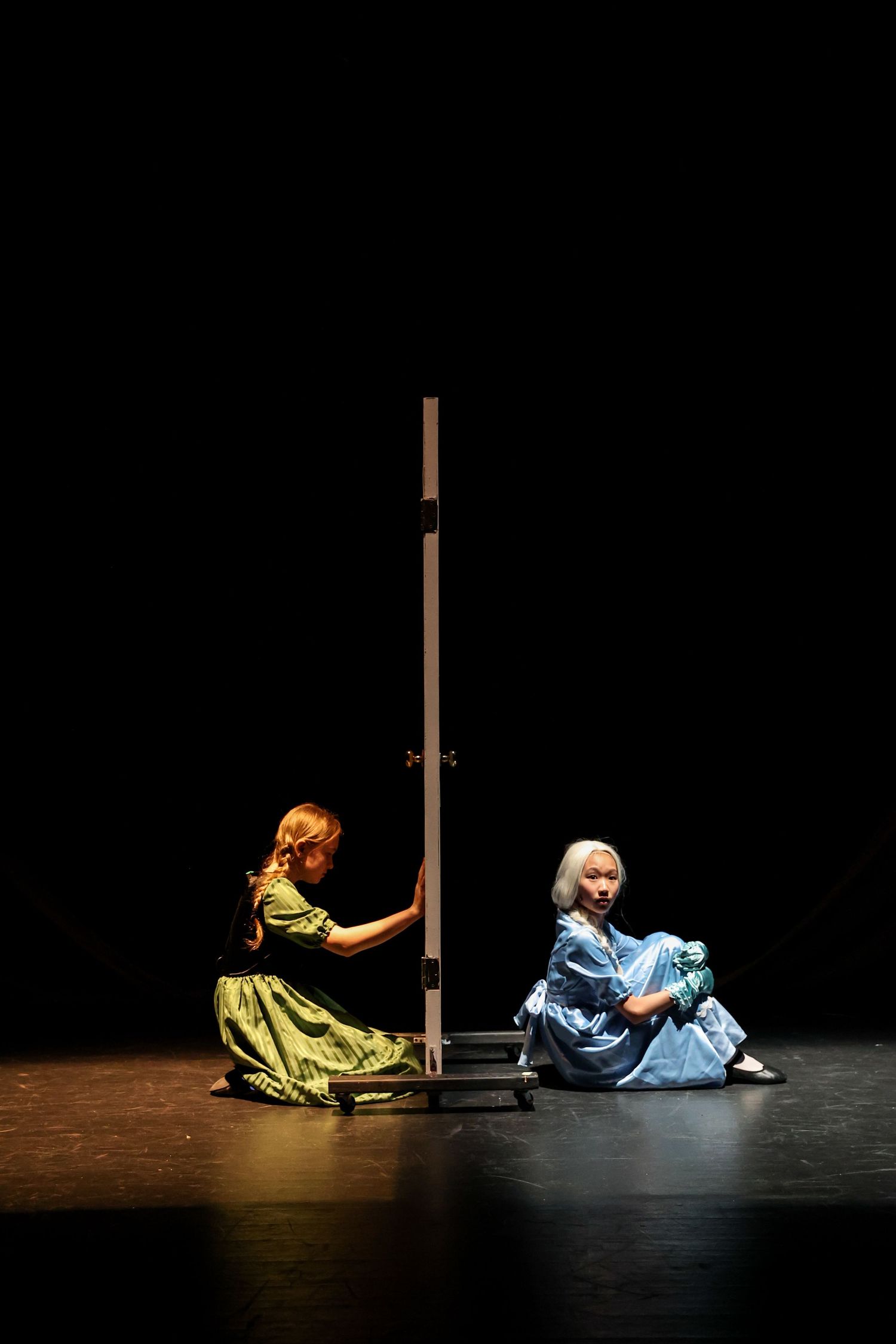 Two performers in period costumes sit on stage floor beside wooden poles in dramatic lighting.