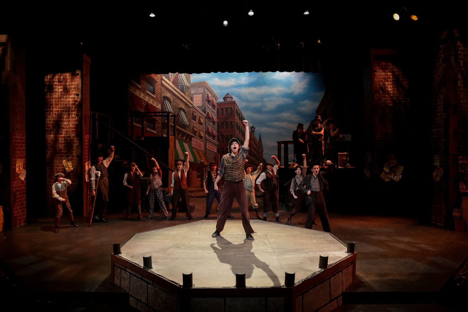 Theatrical performance on a circular stage with urban backdrop and ensemble cast.