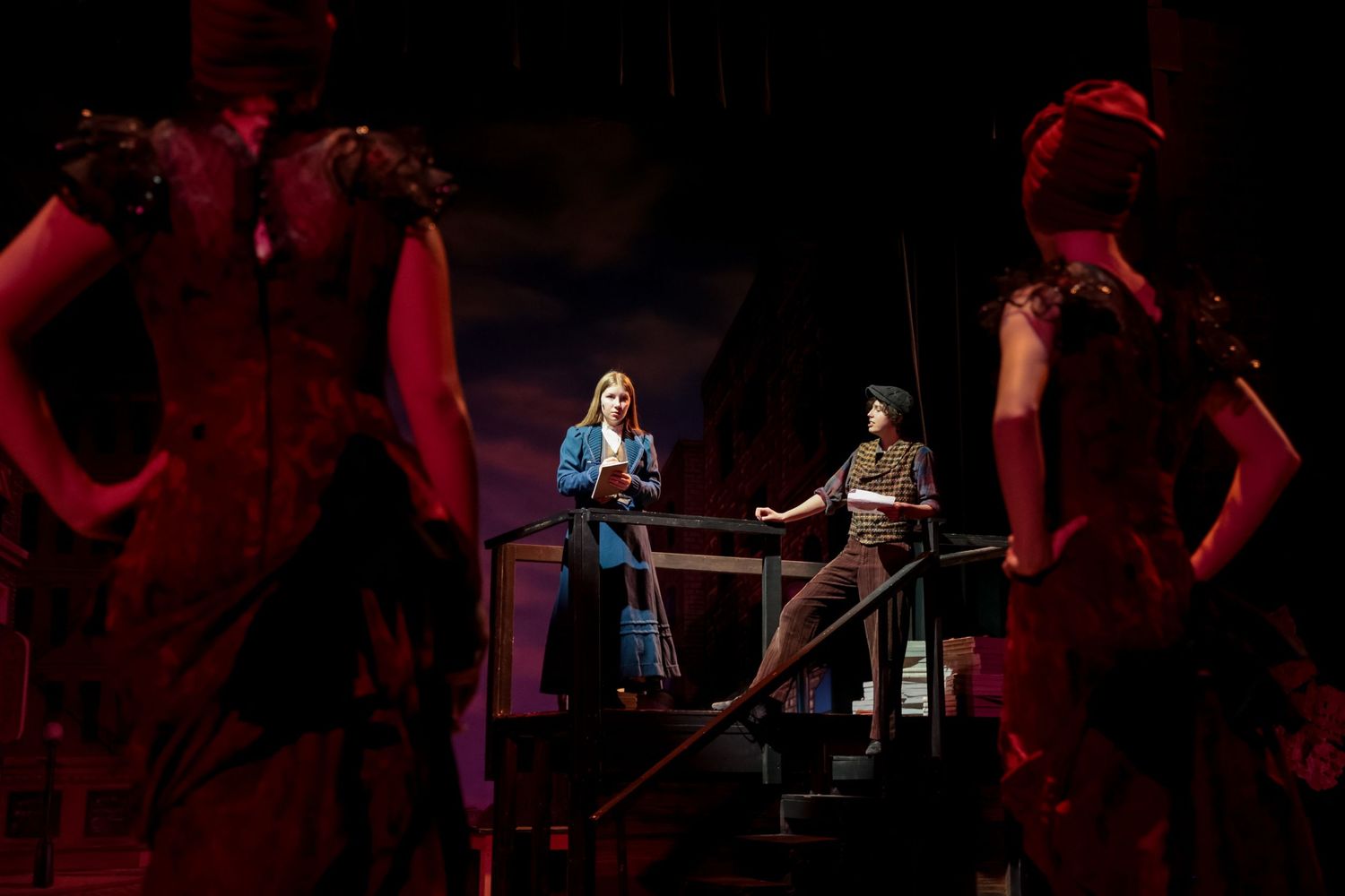 Dramatic stage scene with performers in silhouette against red lighting.