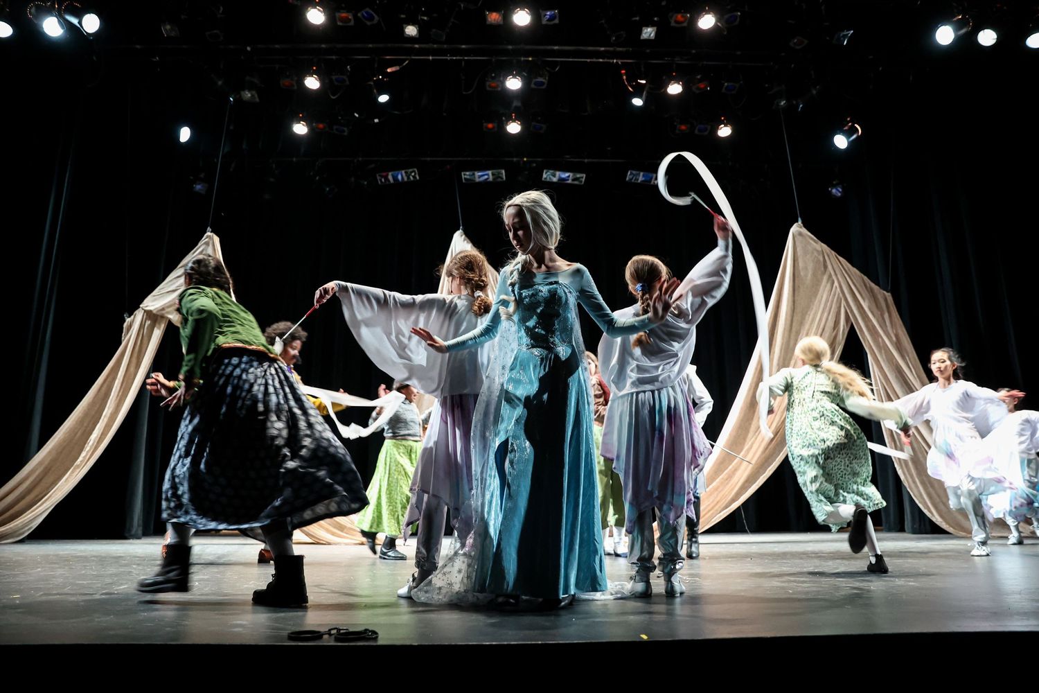 Dance ensemble performs on stage with flowing costumes in bright theatrical lighting.