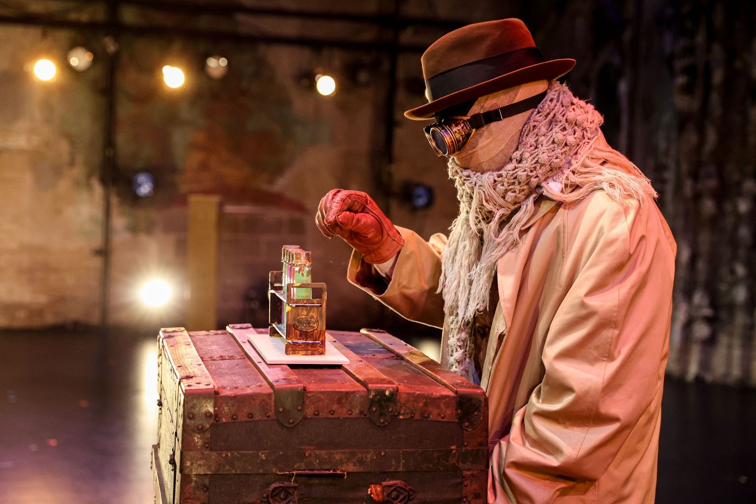 Actor in period costume examining items on an old wooden crate under warm stage lighting.