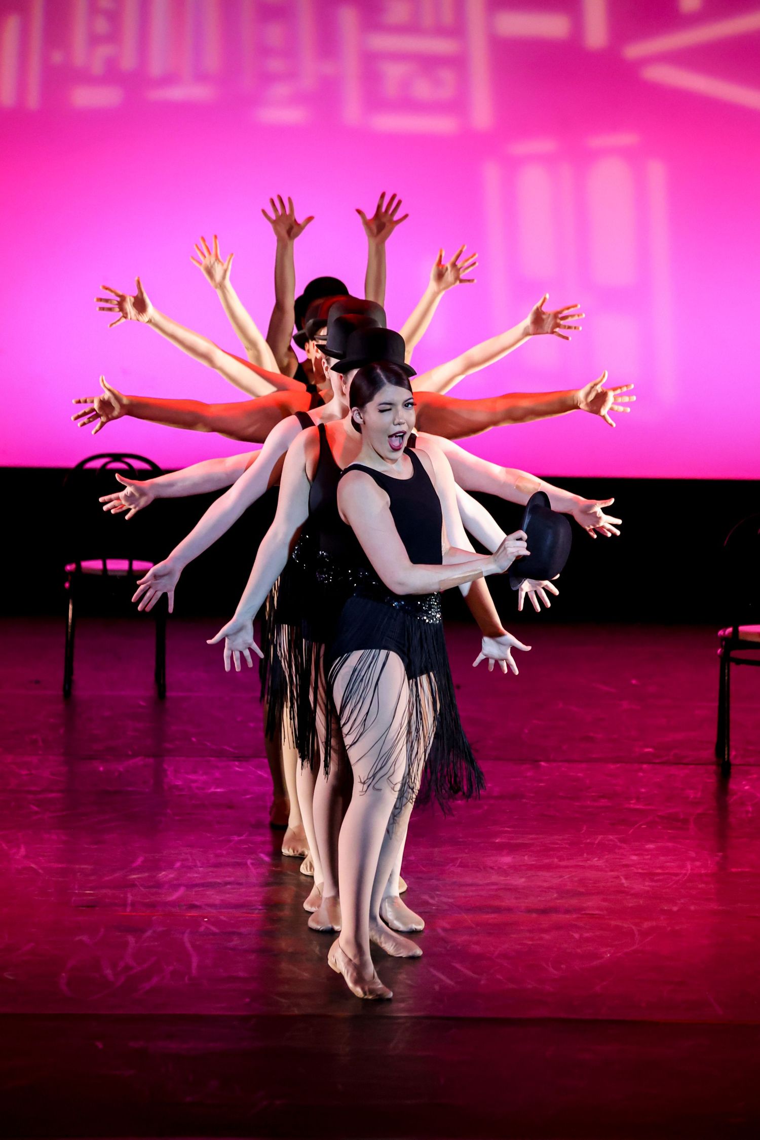 Dancers in formation extend arms outward creating symmetrical patterns against vibrant pink stage lighting.