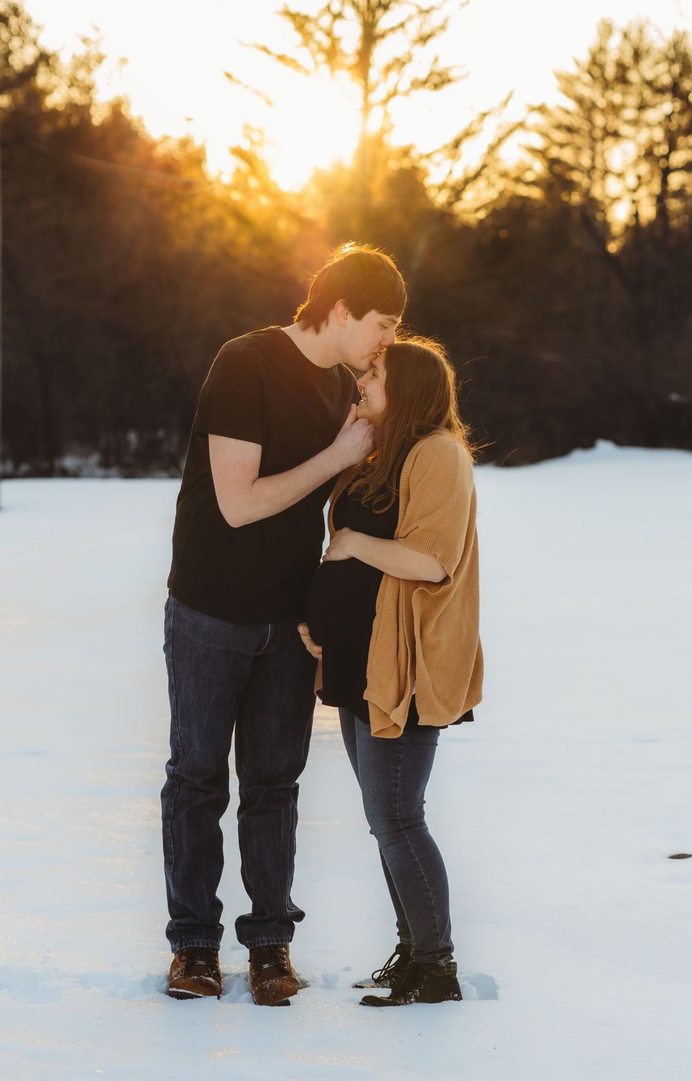 A couple shares an intimate moment in the snow at sunset with golden light filtering through trees.