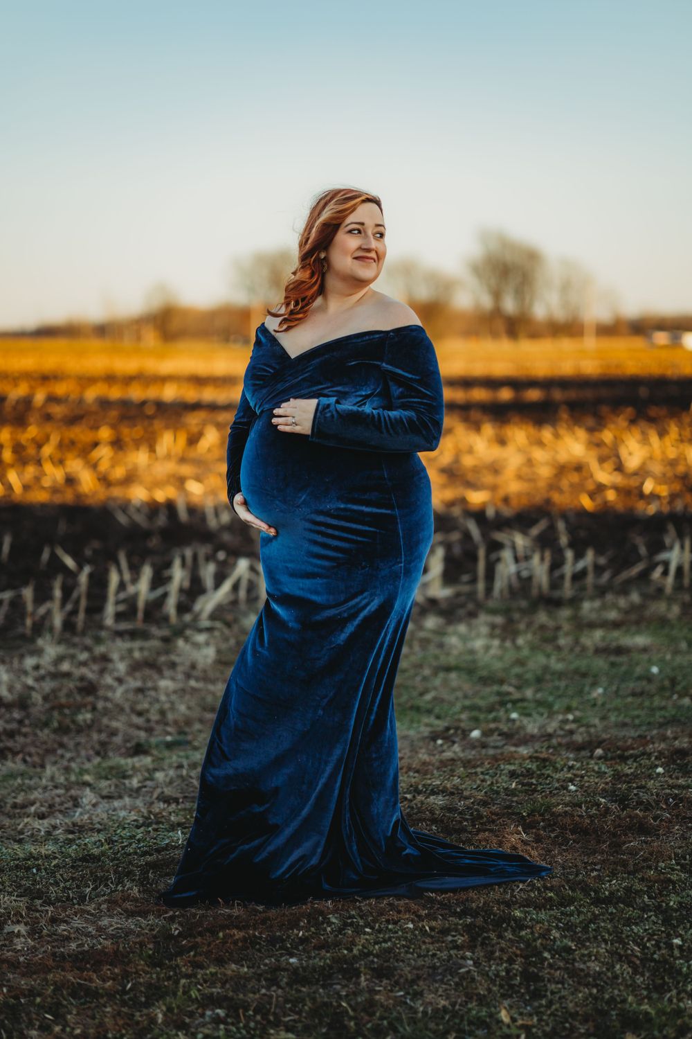 A woman in a royal blue velvet dress poses in a field at golden hour.