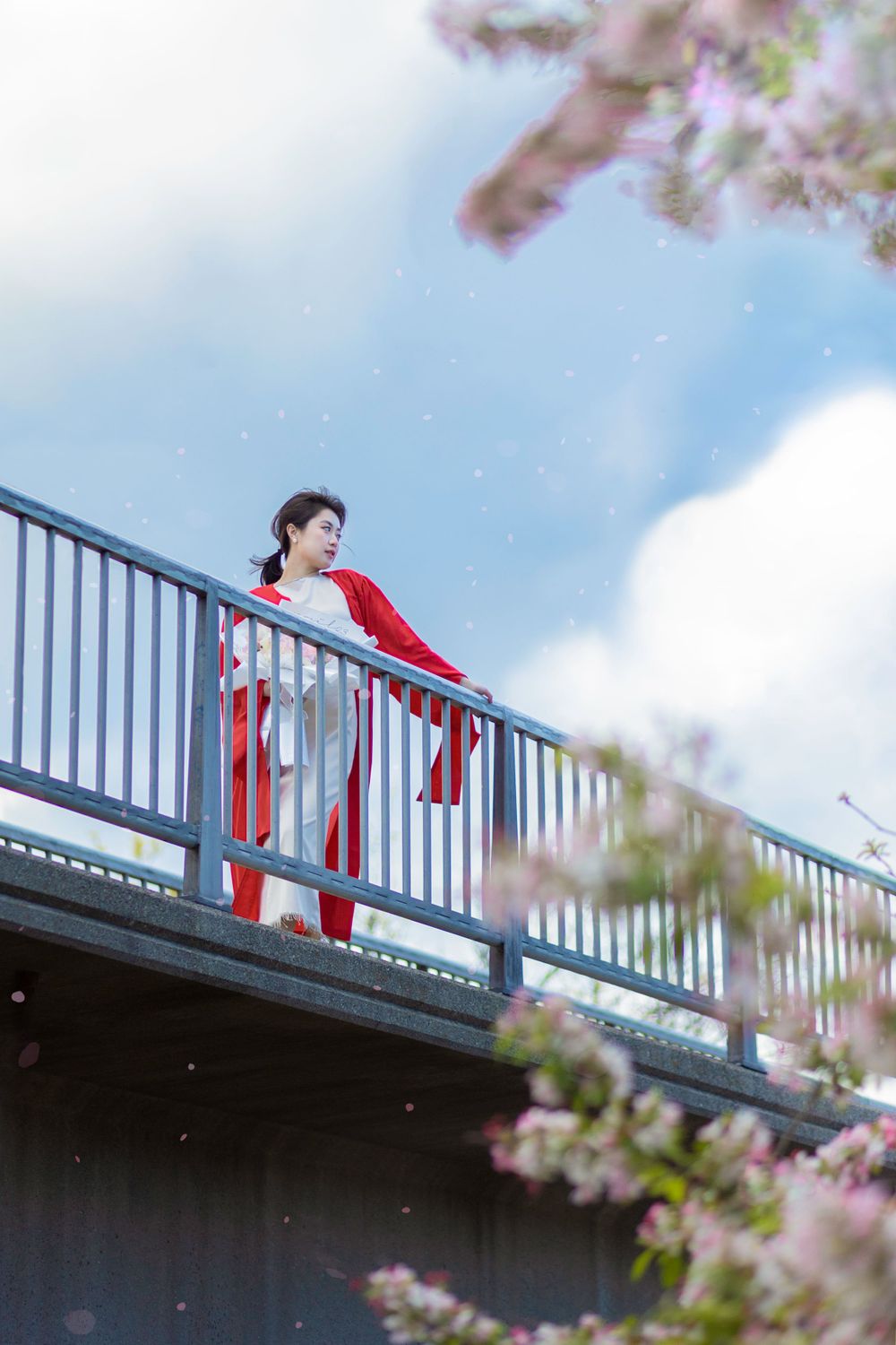 Graduate in academic regalia poses on a railing against blue sky with cherry blossoms in spring.
