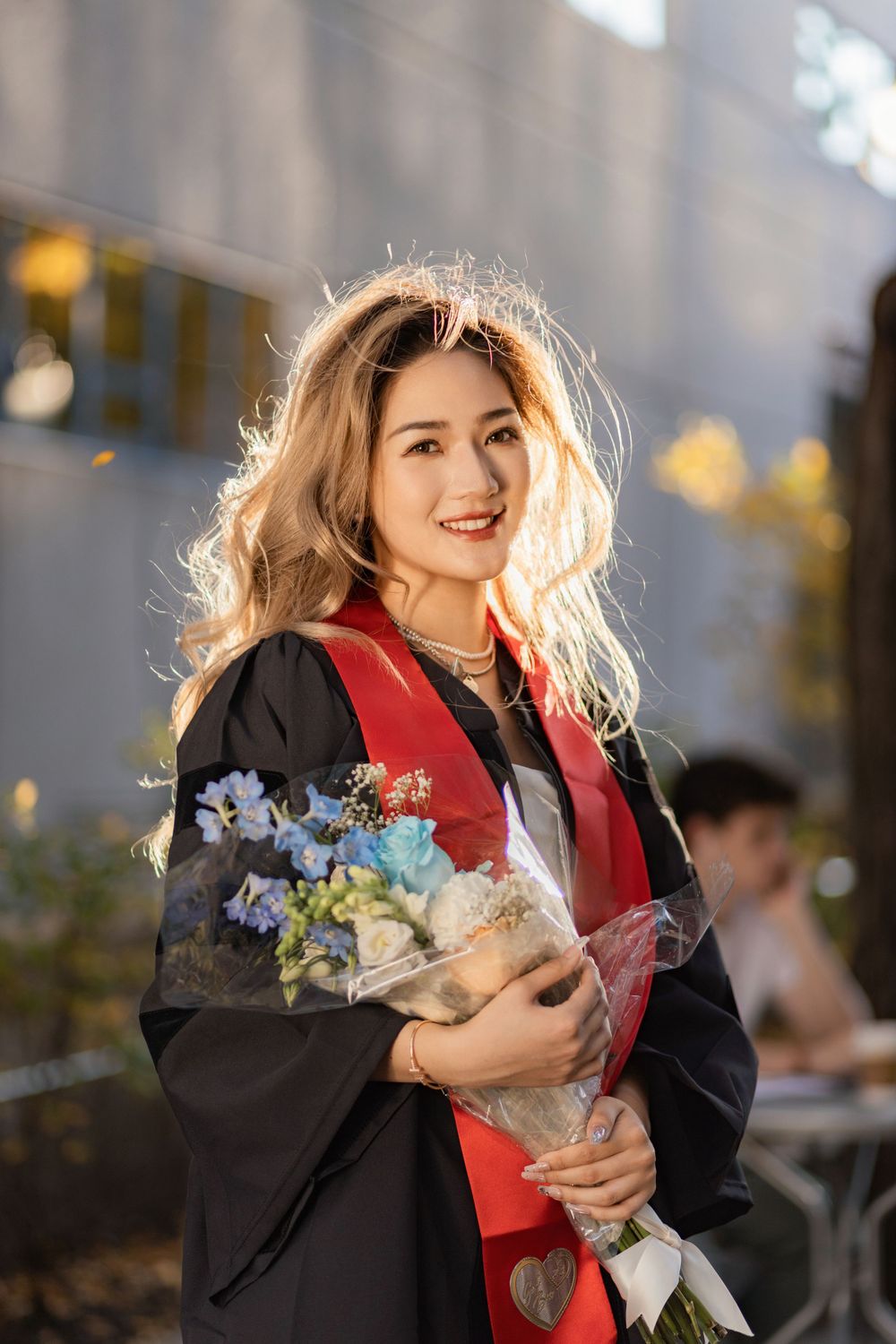 Graduate in black gown and red scarf holds bouquet of flowers during sunset photo session.