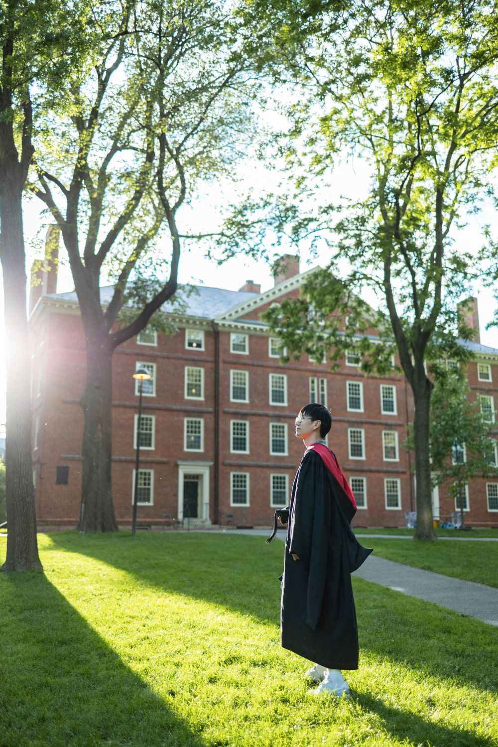 Graduate in cap and gown walks across sunlit campus lawn with brick buildings in background.
