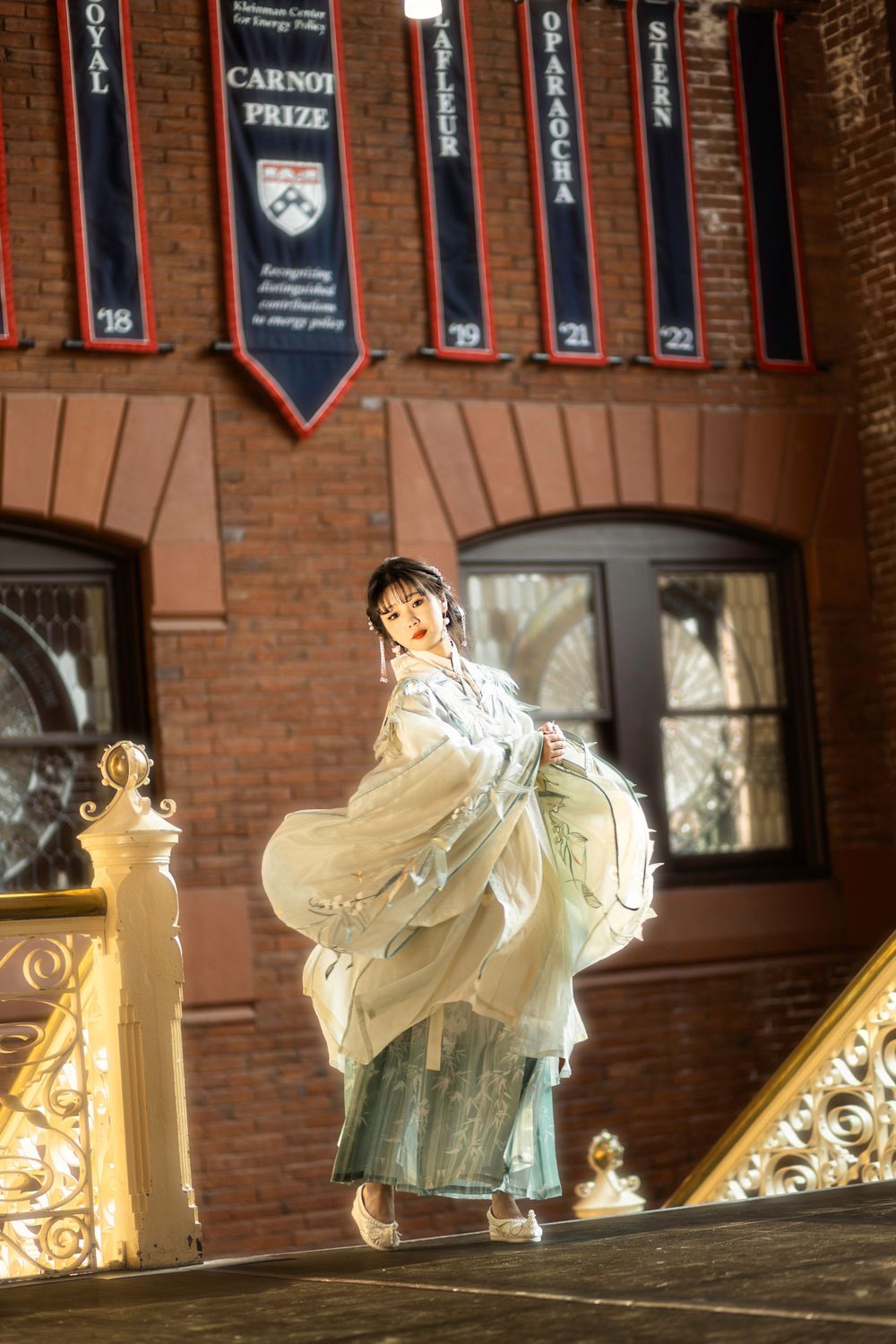 White-clad figures stand along ornate railing in brick building with banners hanging above.