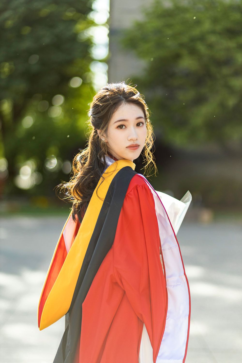 Graduate in red and black academic regalia poses outdoors in natural lighting.