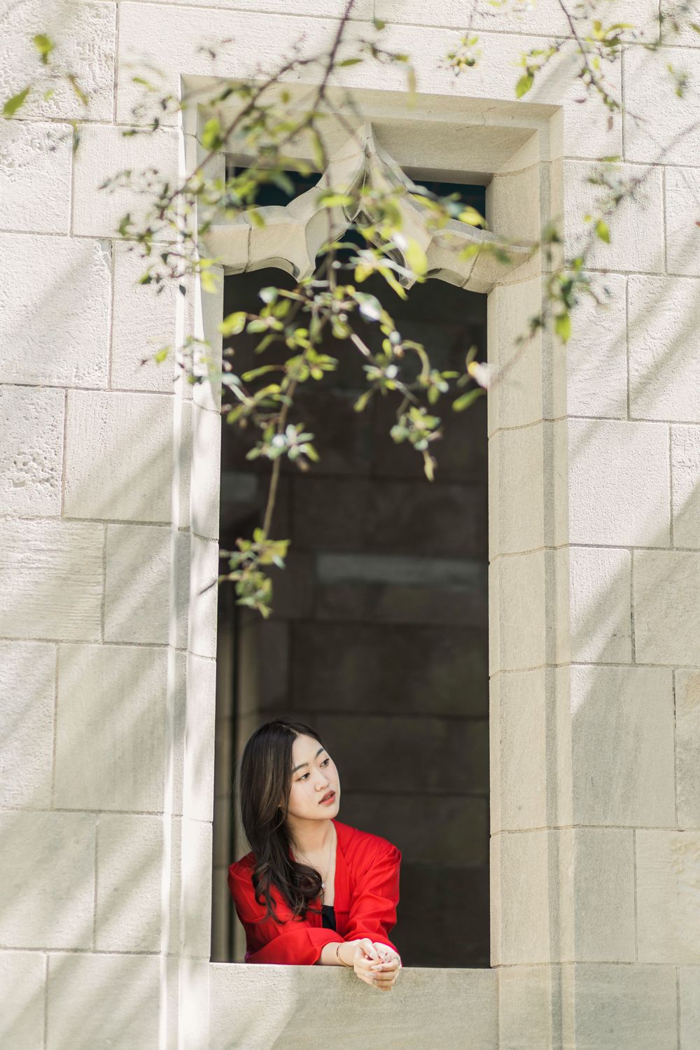 A person in red attire gazes thoughtfully through a framed stone window opening.