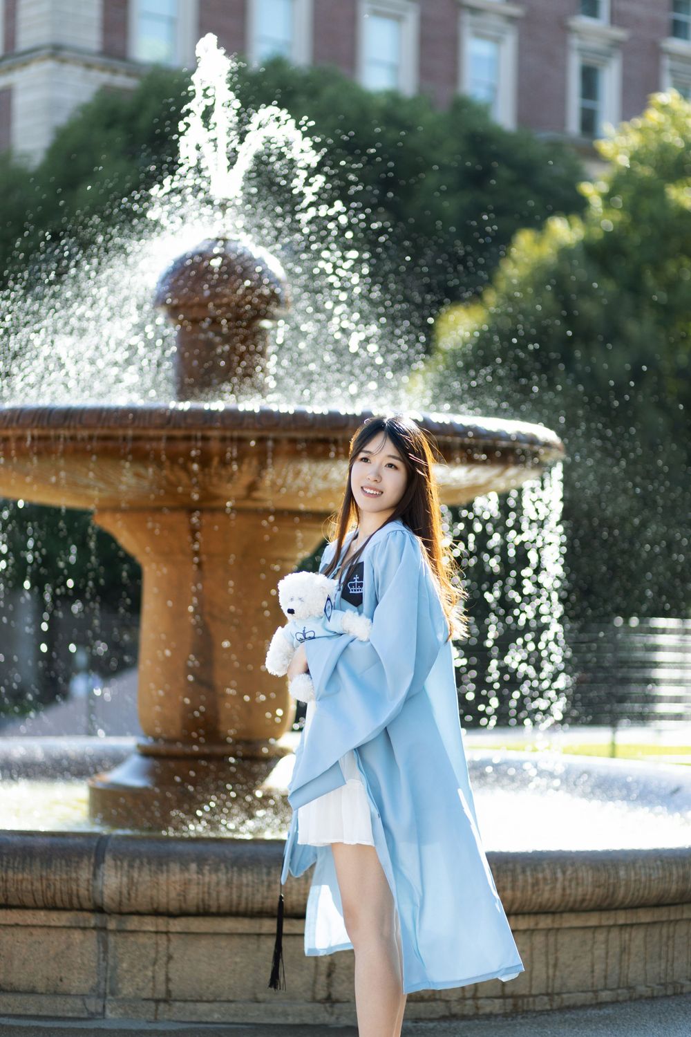 A student in light blue graduation attire poses near a splashing fountain on a sunny day.