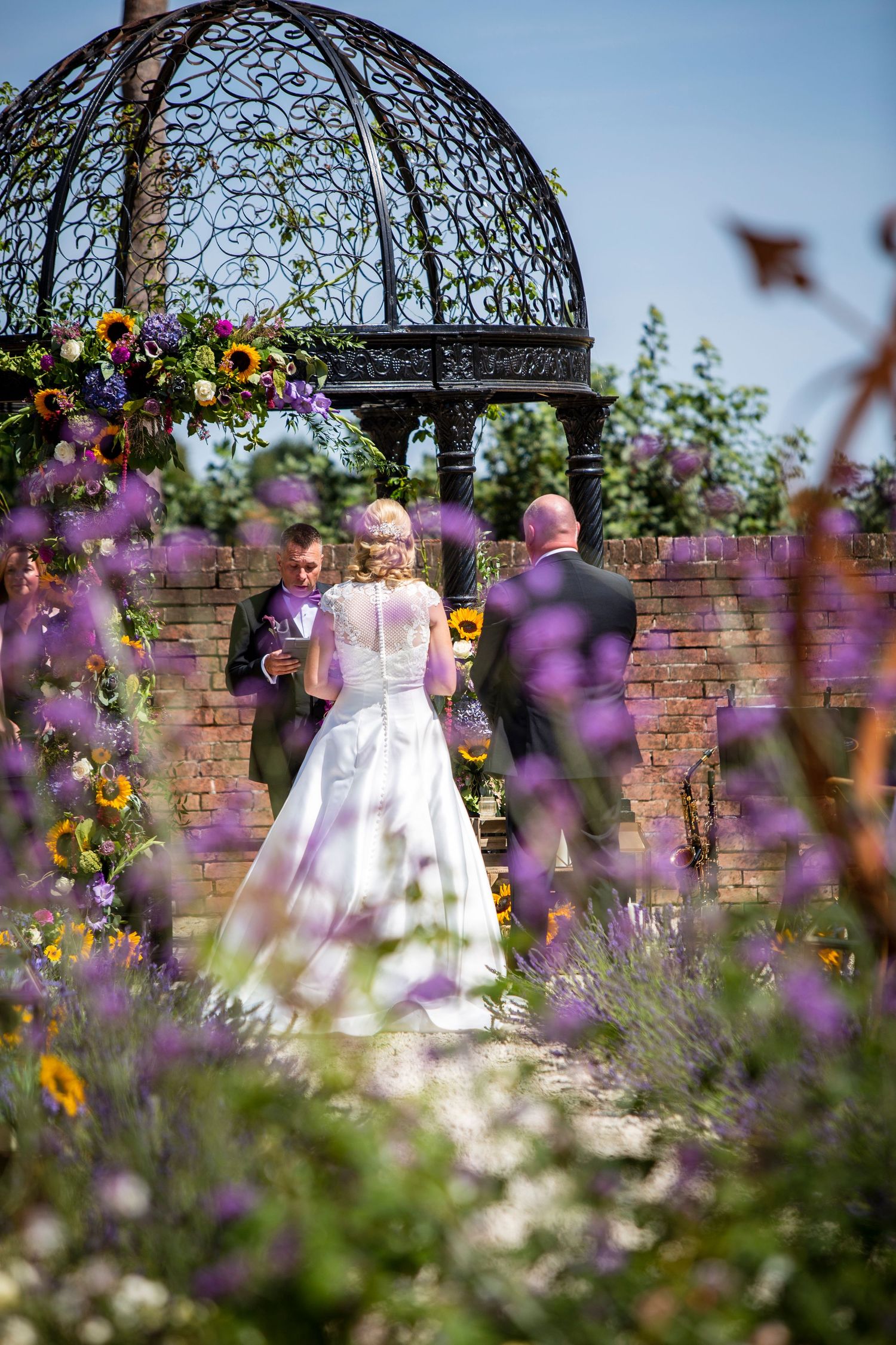Wedding couple under an ornate metal gazebo surrounded by colorful garden flowers.