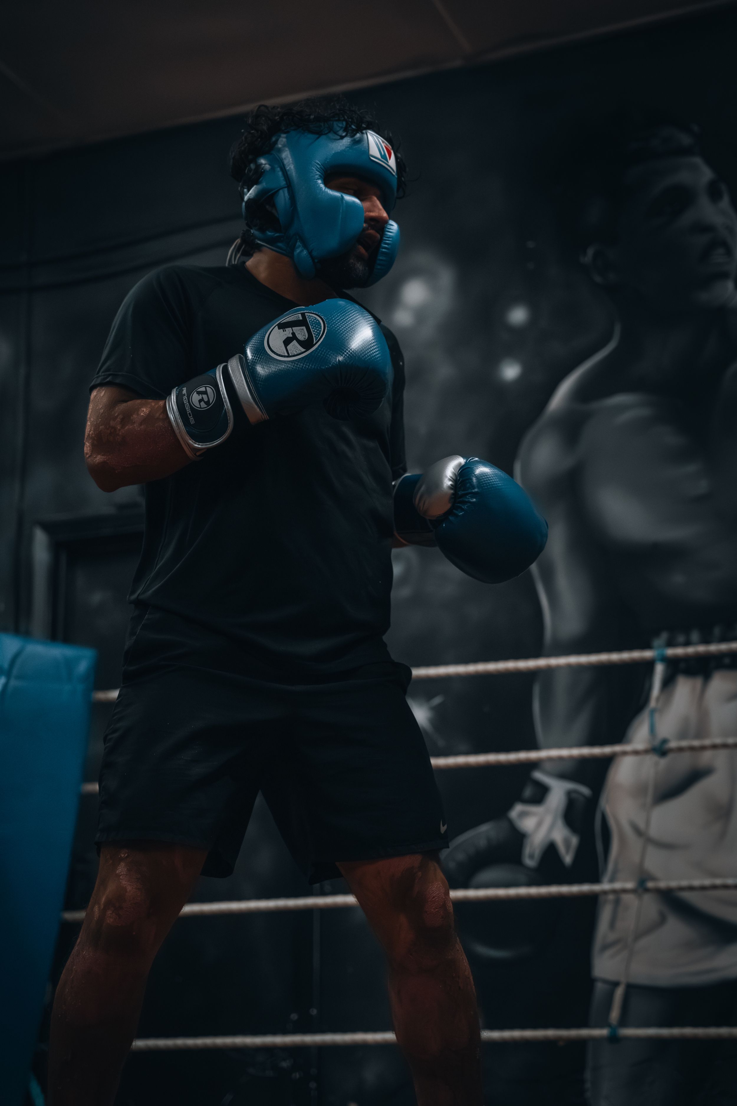 Boxers wearing protective gear train in a moody, dark boxing gym setting.