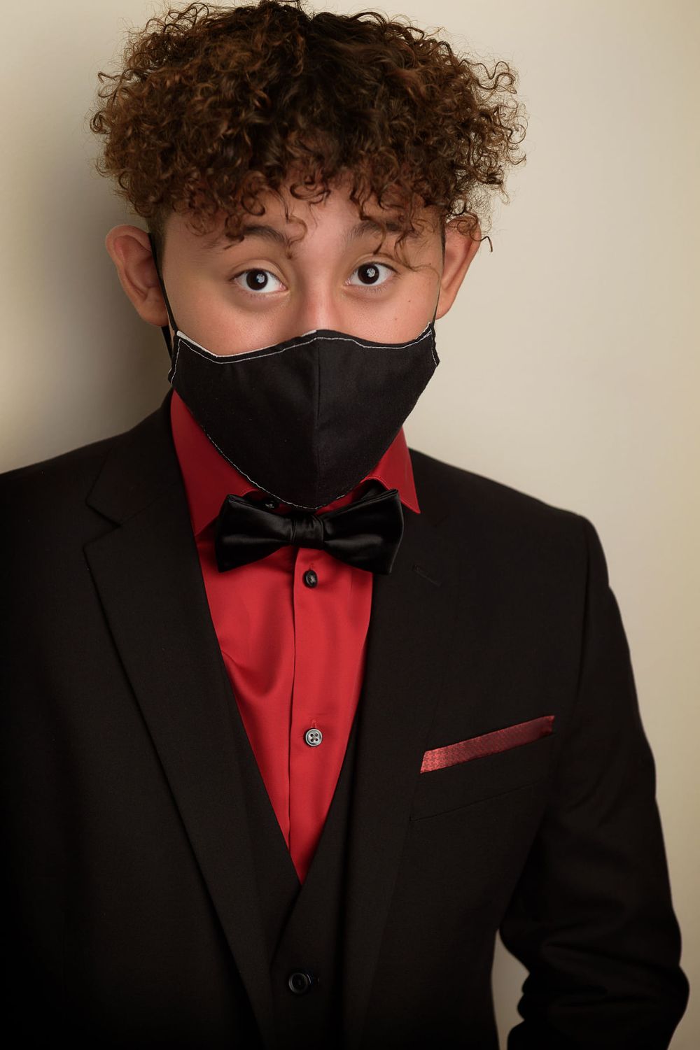 Formal portrait in black suit with red shirt, black bow tie and face mask against light background.