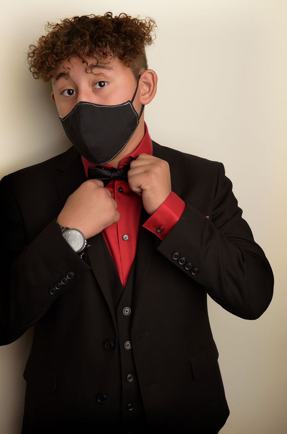 Studio portrait showing black suit, red shirt and protective mask while adjusting collar.