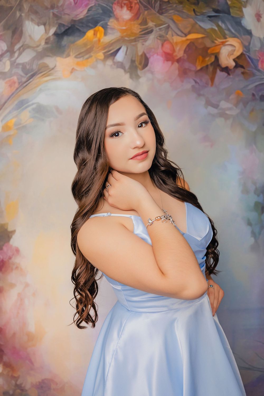 A portrait photo against a painted floral backdrop featuring someone in a light blue dress with long wavy hair.