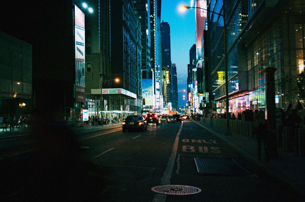 Nighttime city street scene with blue-tinted lighting and illuminated buildings.