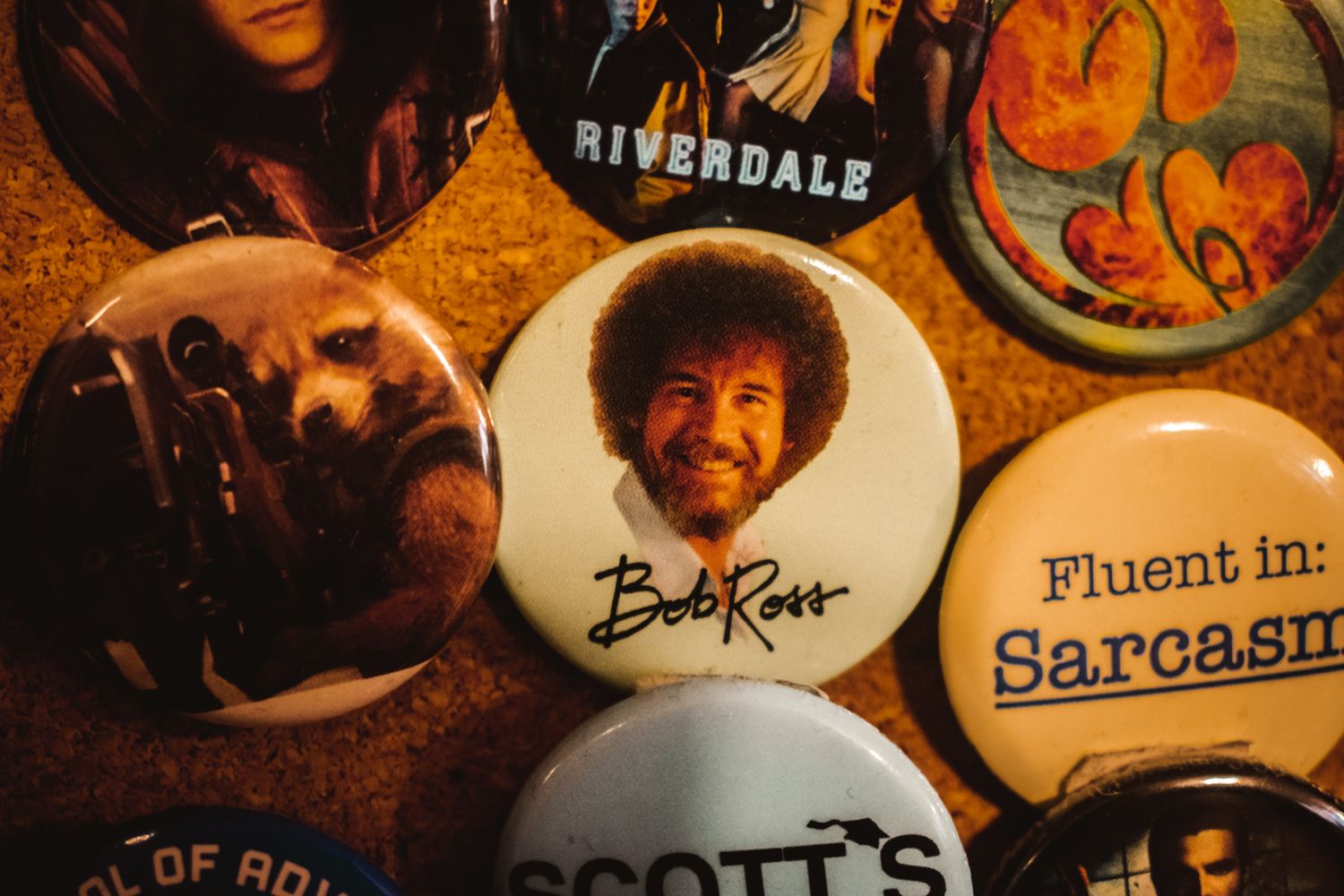Collection of pop culture pins and buttons including Riverdale and Bob Ross merchandise.