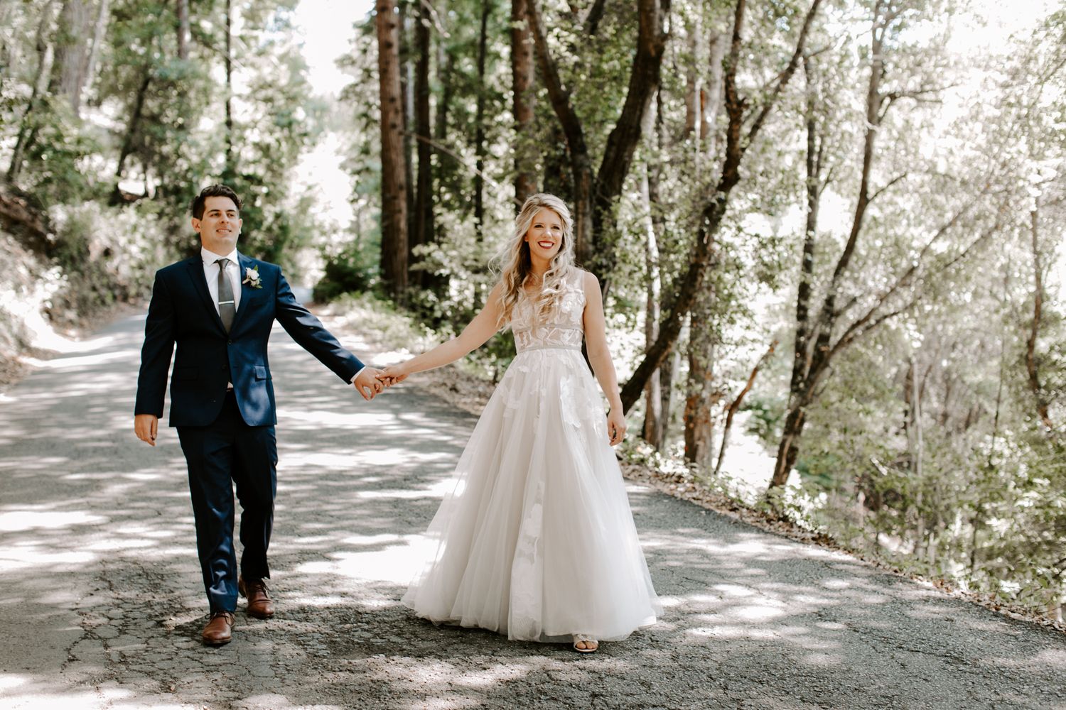 A newlywed couple walks hand in hand along a tree-lined path in a forest setting.