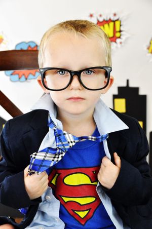 A young child in glasses wearing a Superman shirt and blazer poses against a comic-themed backdrop.