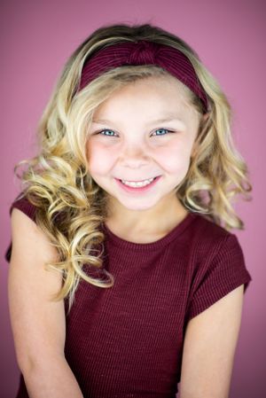 Studio portrait against a pink background featuring blonde curls and a burgundy top.