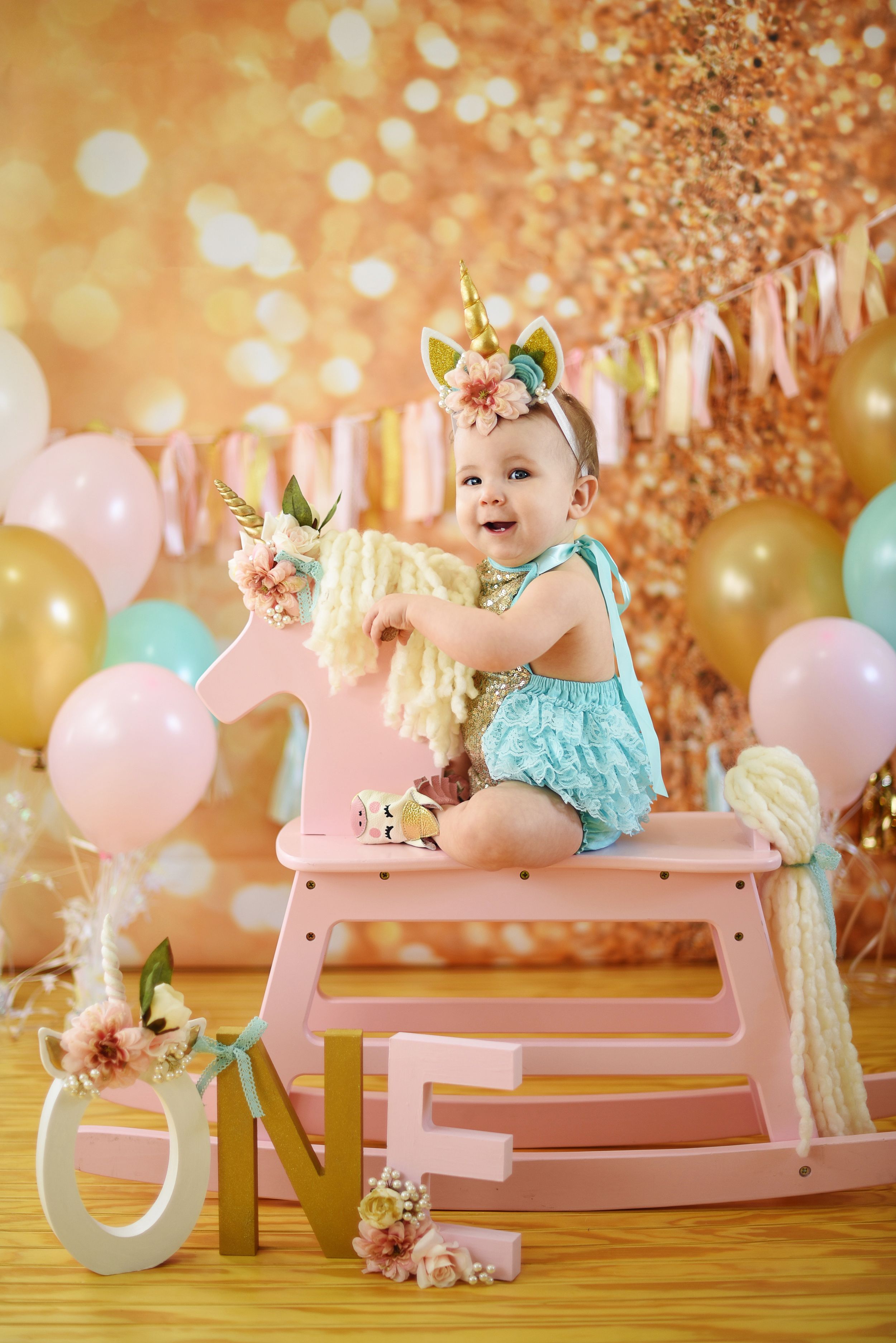 Baby in teal outfit celebrates first birthday with pink and gold party decorations.