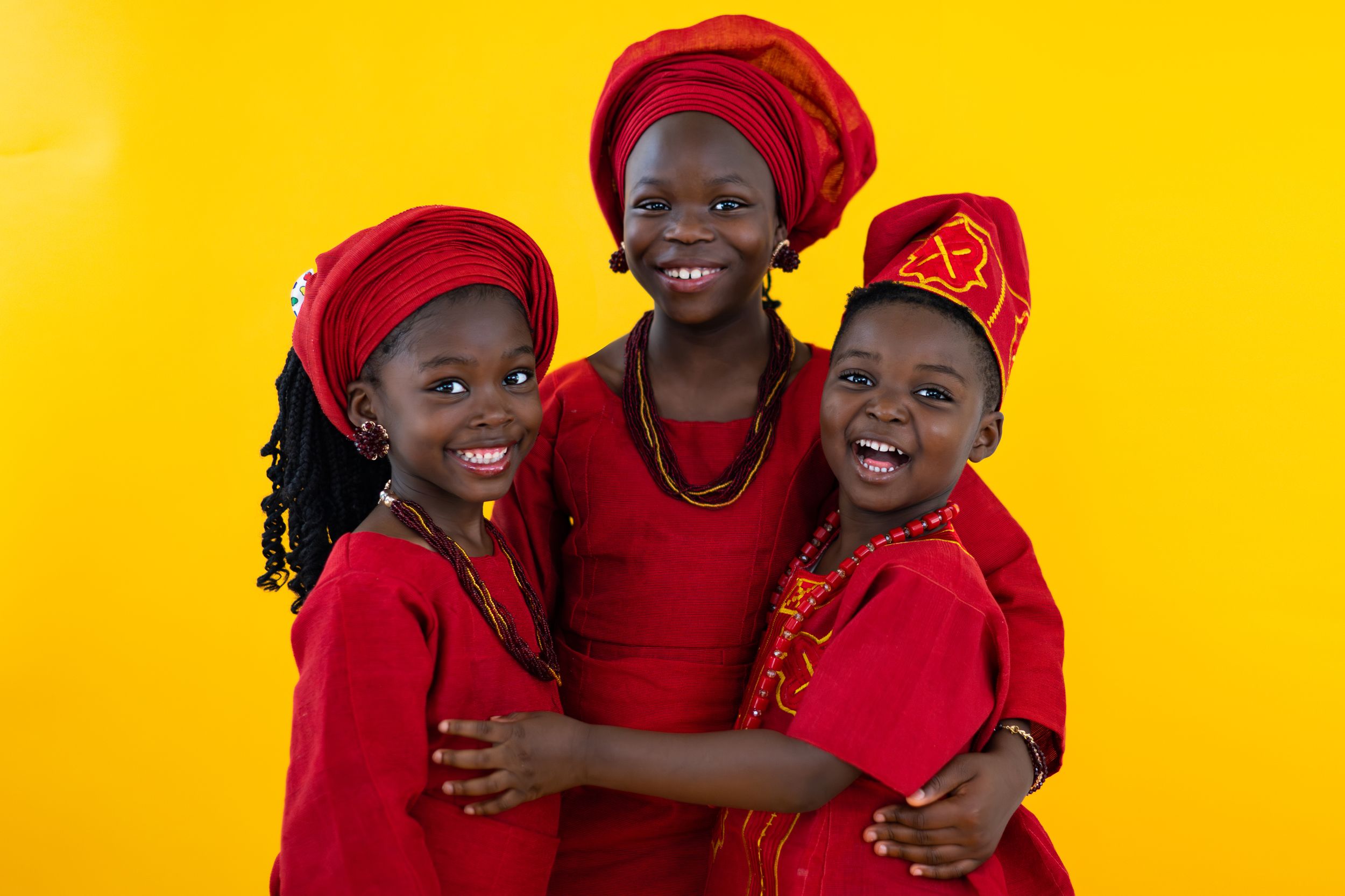 Three people in bright red outfits and traditional head wraps pose joyfully against a yellow background.