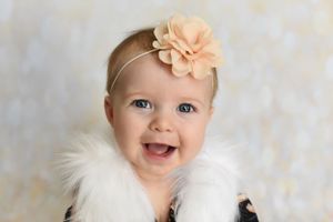 A joyful baby wearing a peach flower headband and white fur wrap.