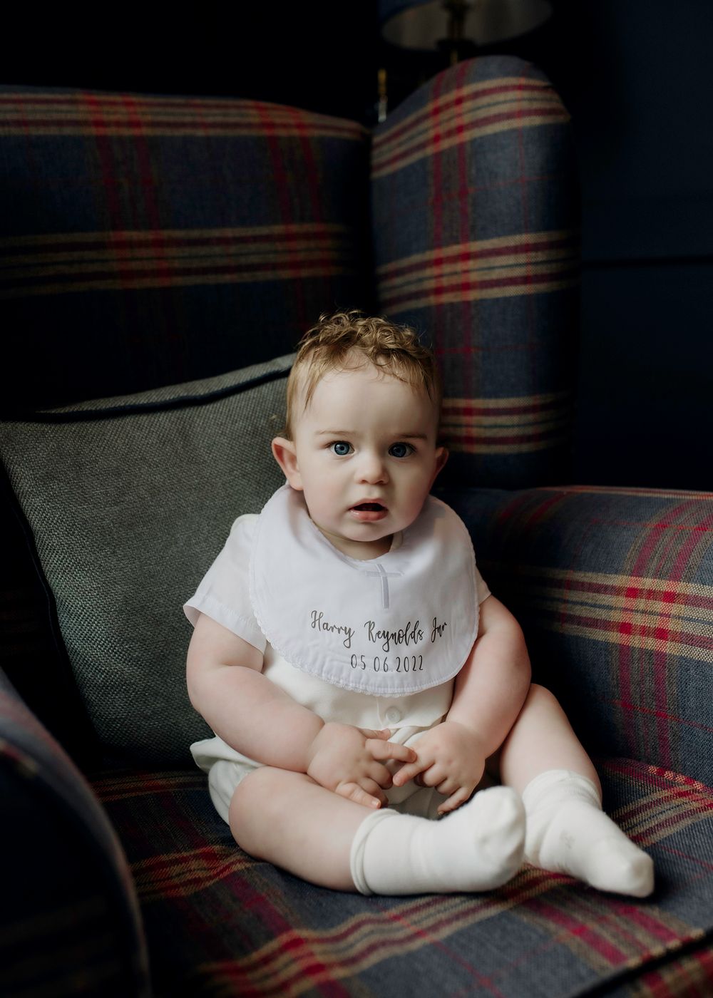 A baby sits on a plaid armchair, wearing a personalized white bib with the name "Harry Reynolds Jr." and a date, surrounded by soft lighting