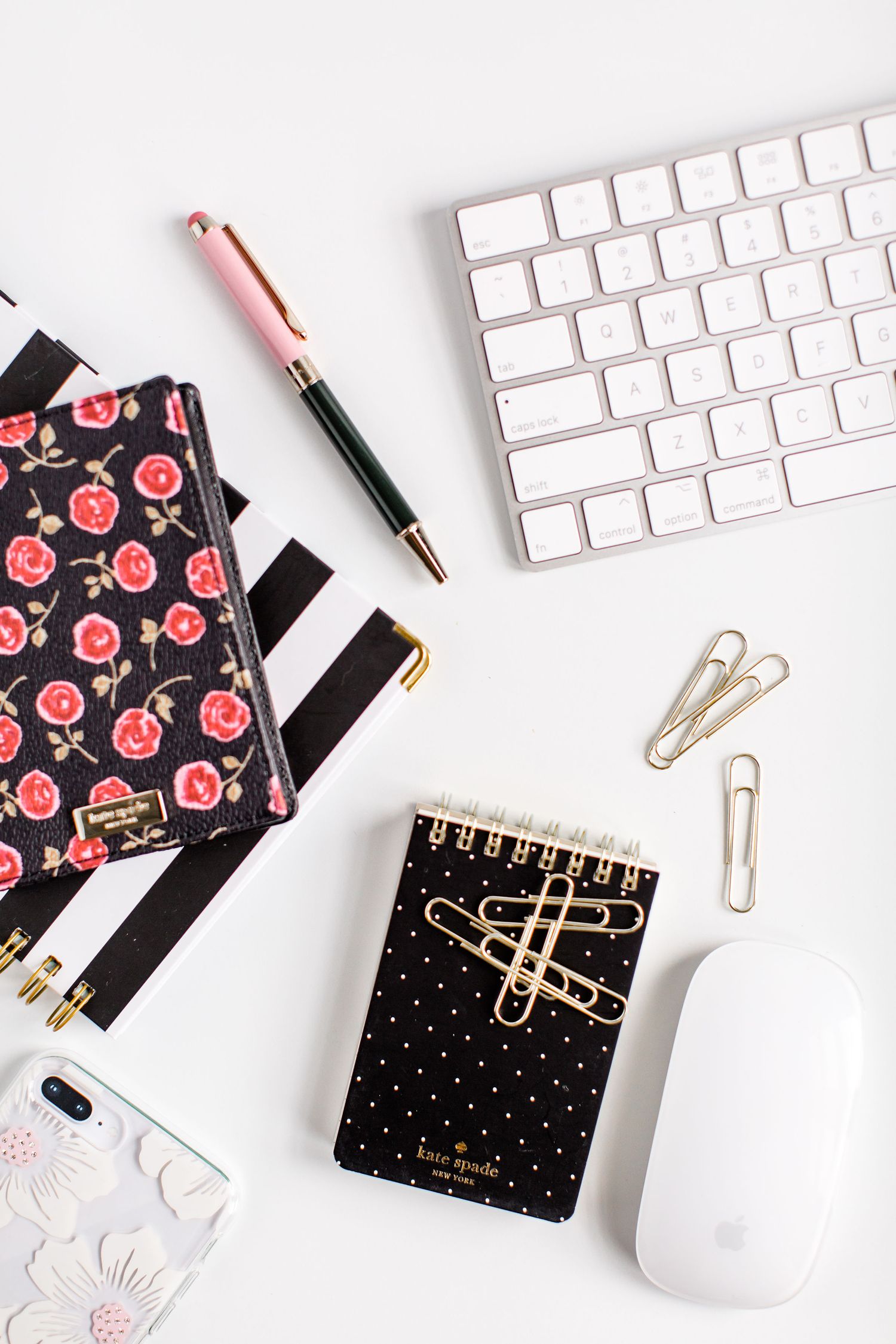 Flatlay of desk accessories including floral notebook, keyboard, mouse and gold paper clips.