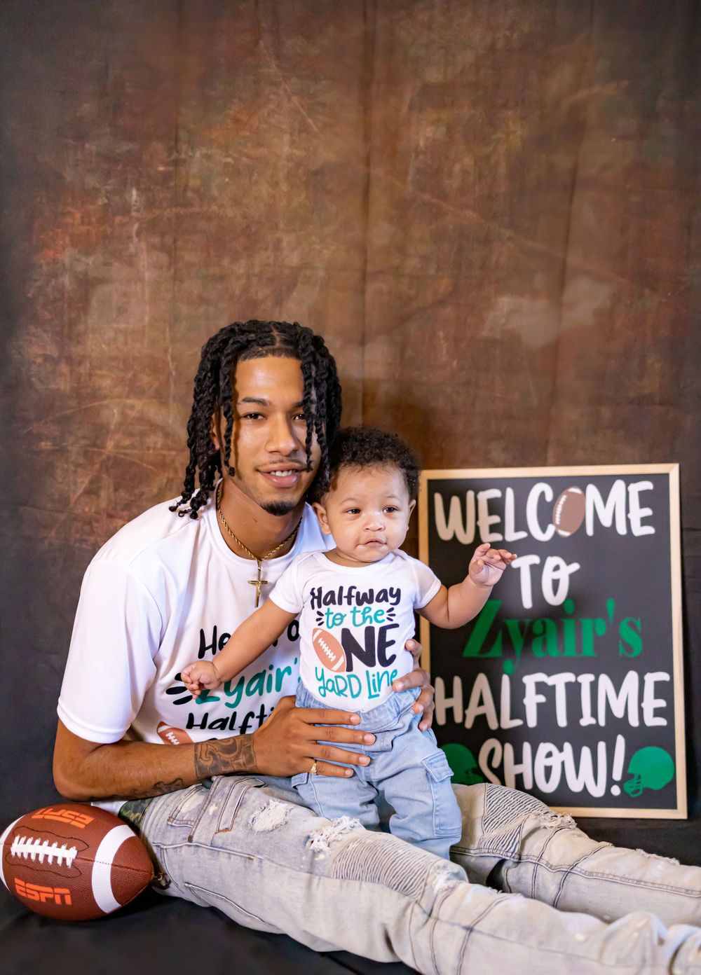 Parent and baby pose with football and chalkboard sign for halftime show themed photo.