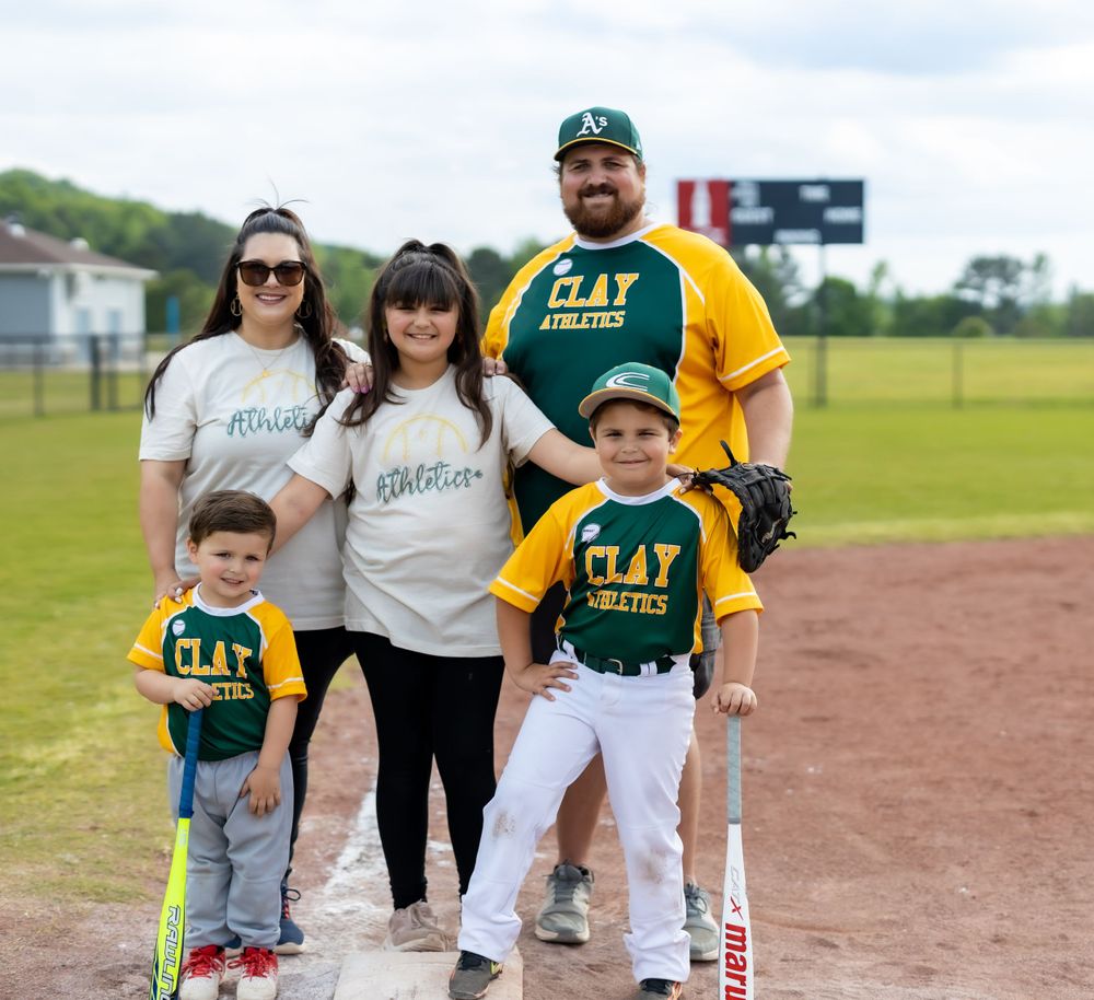 Family in Clay Athletics uniforms poses together on baseball field with sports equipment.