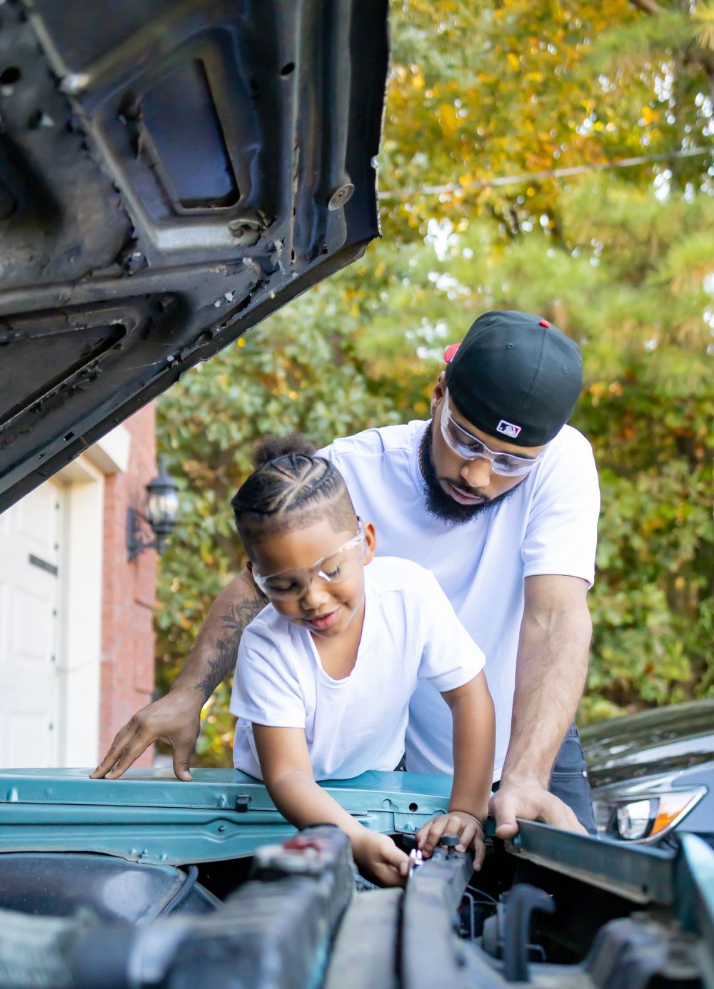 A person in a white t-shirt inspects under the hood of a car with assistance.