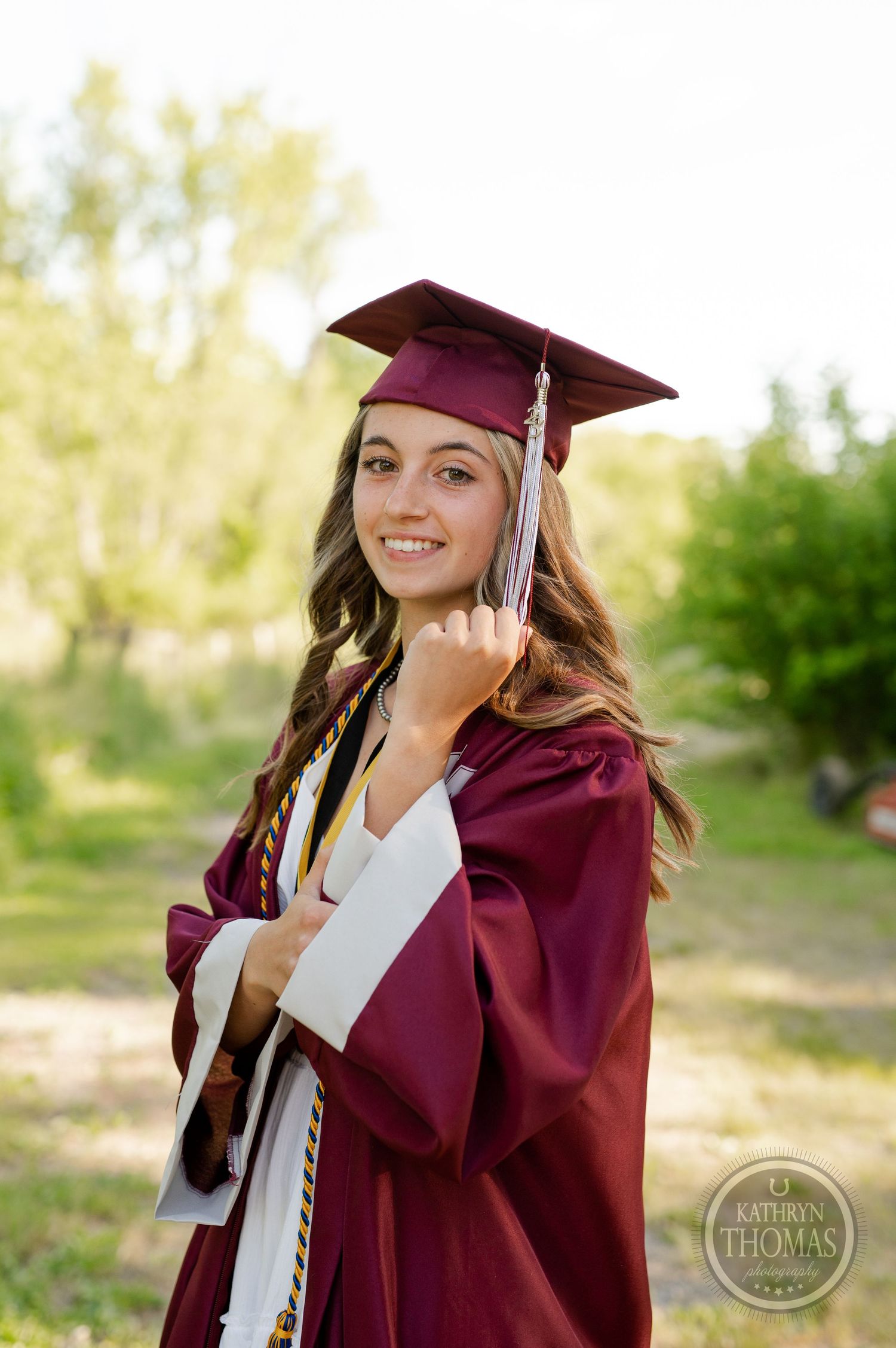 Graduate in maroon cap and gown poses outdoors with diploma against natural backdrop.